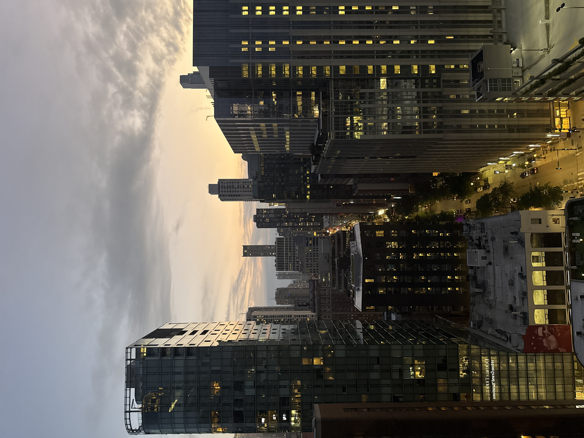 The image captures a city skyline at dusk, likely Chicago, United States of America, viewed from a high vantage point. The sky is a gradient of pale blue and soft orange, with scattered clouds creating texture. A prominent, tall, glass-encased building with a curved corner dominates the left side of the frame, its windows reflecting the twilight. To its right, a dense collection of other skyscrapers of varying heights and architectural styles fill the midground and background. Many of these buildings have illuminated windows, suggesting occupancy. A street is visible below, lined with trees and illuminated by streetlights, with a few cars and possibly pedestrians present. The overall impression is one of urban life transitioning from day to night, characterized by modern architecture and a soft, diffused light.