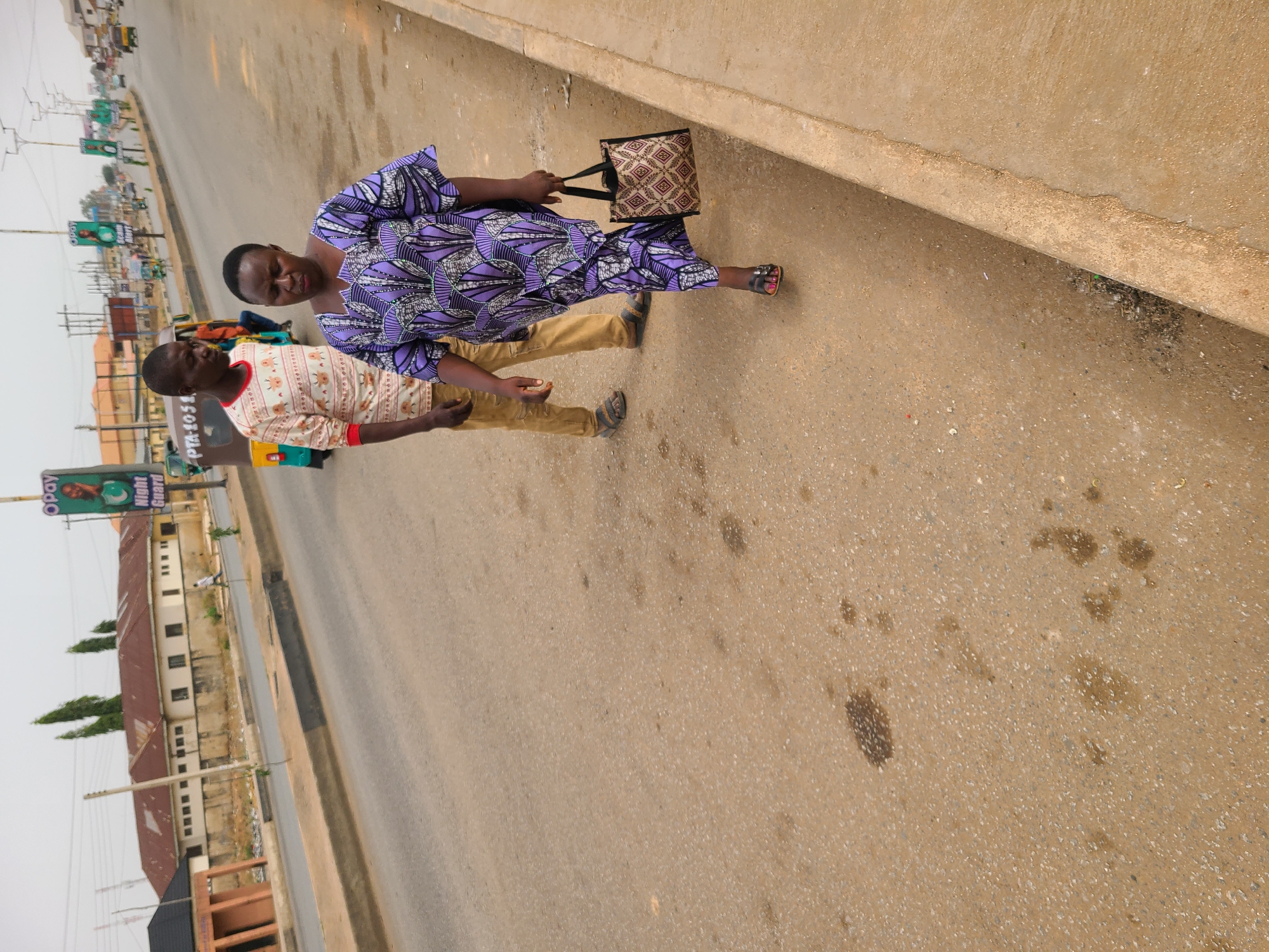 Two people are walking on a paved road with a curb in Jos, Nigeria. One person is wearing a purple patterned dress and carrying a brown purse. The second person is wearing a white and red patterned shirt and khaki pants. Buildings and signs are visible in the background.