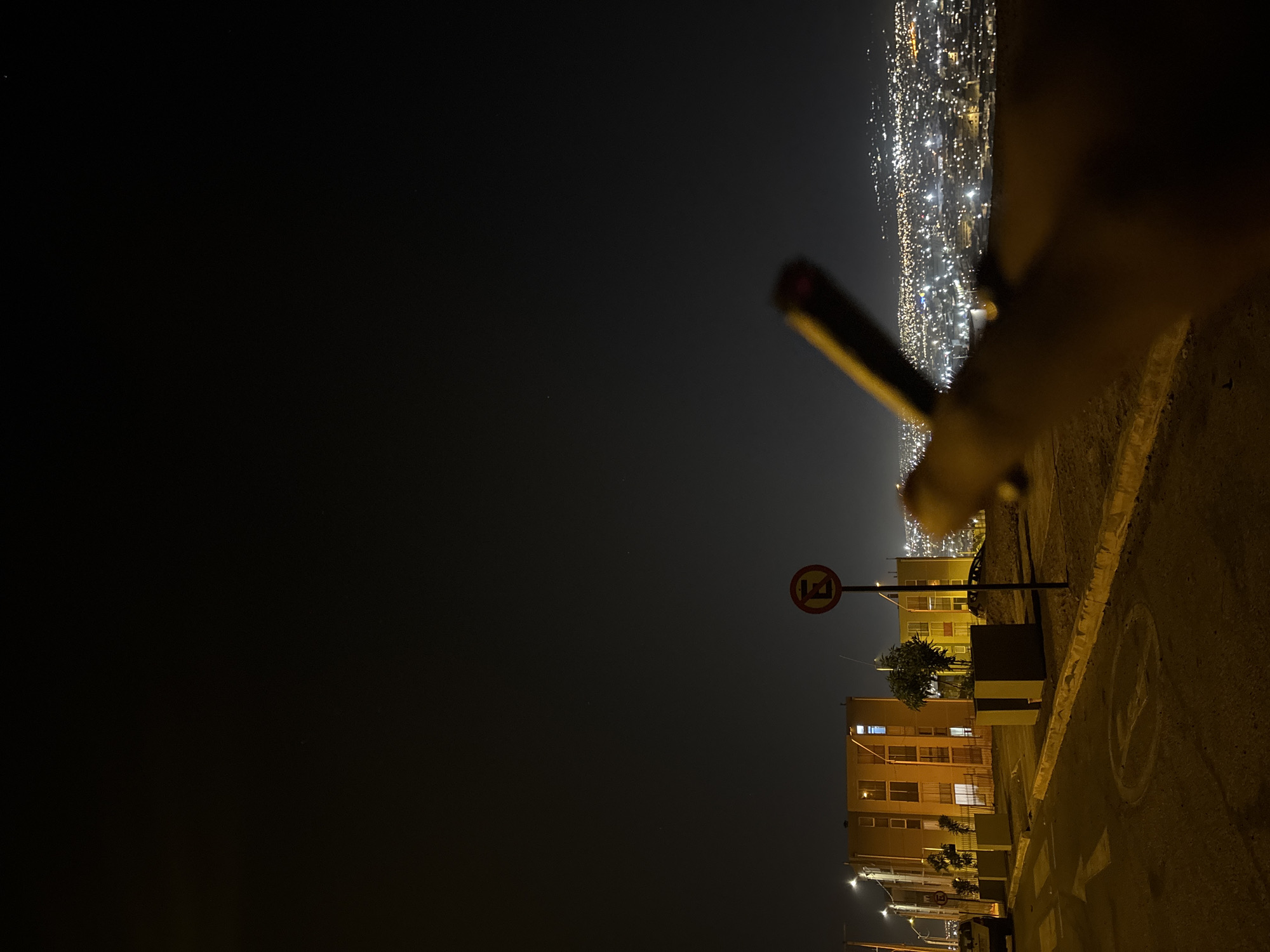 The image depicts a night scene in Arica, Chile. A portion of a hand holding a thin, dark object is visible in the foreground on the right. Below the hand, there is a concrete wall next to an illuminated street. A "no parking" sign stands near a building with lit windows. The background reveals a city view with numerous lights dotting the landscape. The sky above the city is dark. The scene is illuminated by artificial lights.