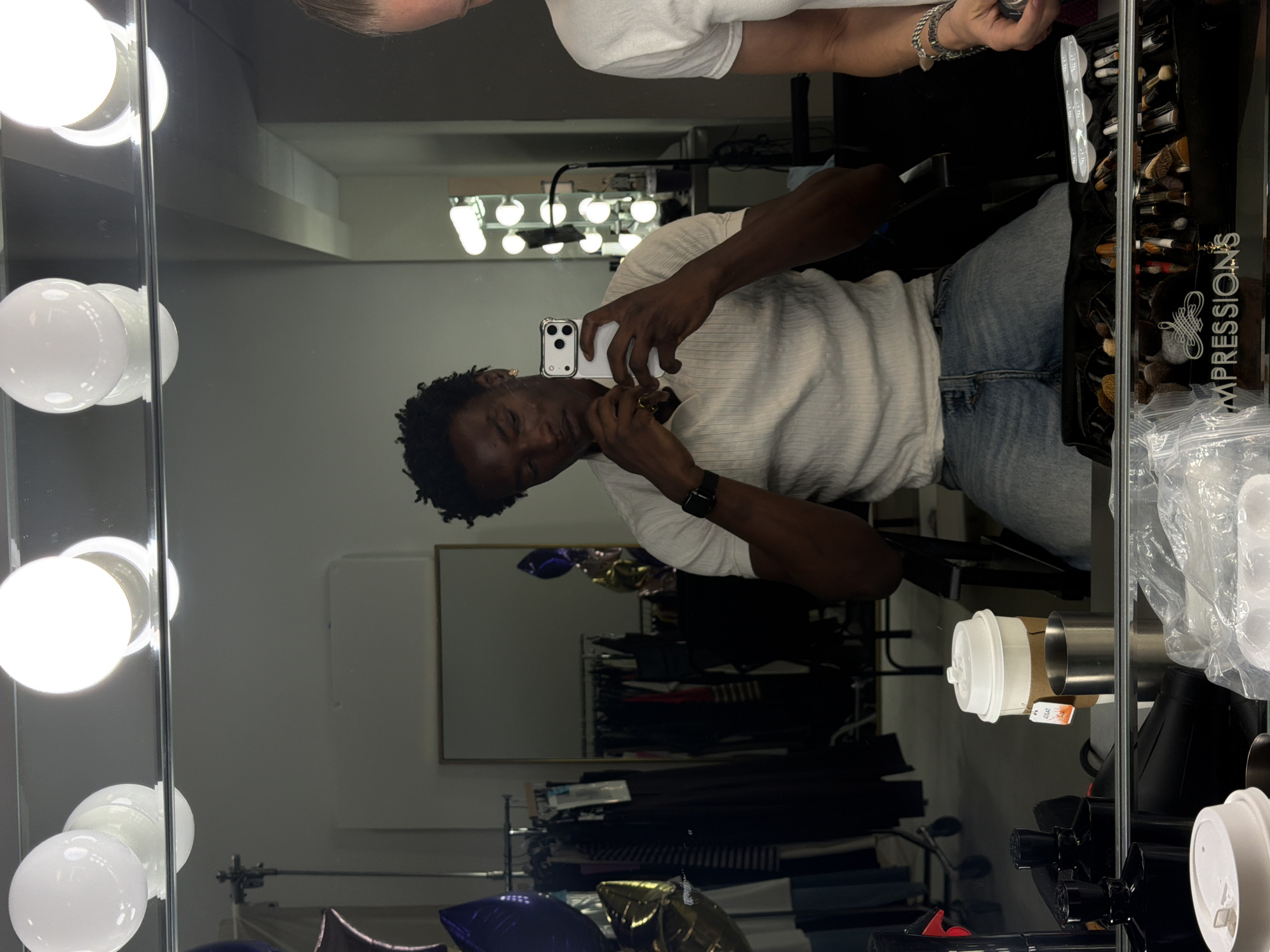 A Black man is seated in a chair, taking a selfie in a mirror that reflects a dressing room. The dressing room is illuminated by vanity lights above the mirror. The man is wearing a light-colored polo shirt and blue jeans. He is holding a white smartphone in his right hand, and his left hand is resting on his chin. A watch is visible on his left wrist. In the reflection, a rack of clothing and some balloons are visible. To the right of the man, in the reflection, another person wearing a white shirt is partially visible. In the foreground of the image, on a surface below the mirror, a white disposable coffee cup with a lid and a brown sleeve, a silver metal cup, and various makeup brushes in a black case are visible. The location is identified as New Albany, United States of America.