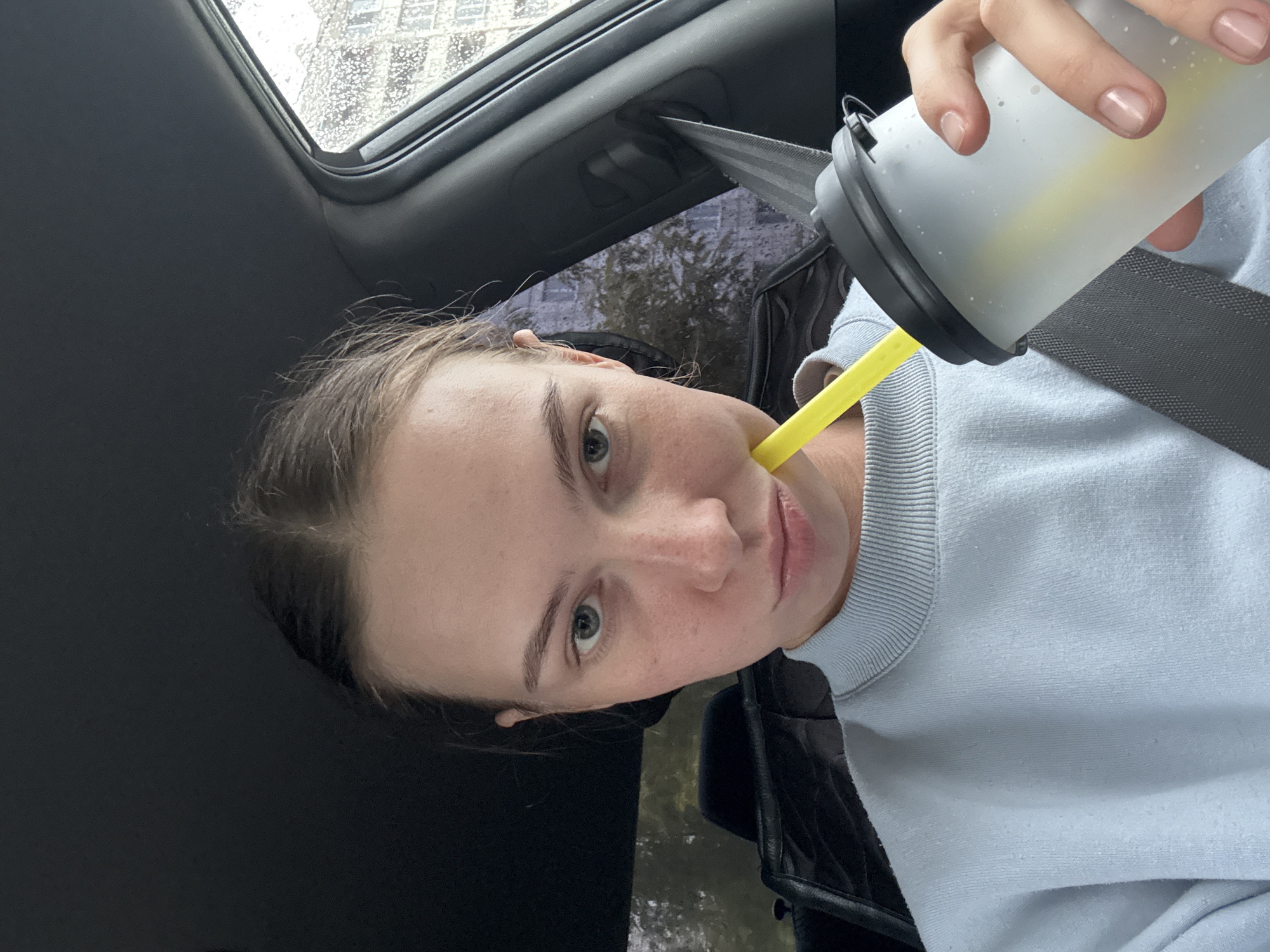 A young Caucasian woman with light skin and freckles is taking a selfie in the driver's seat of a car. She has light brown hair pulled back and is wearing a light blue sweatshirt. She is holding a frosted white reusable cup with a yellow straw and appears to be drinking from it. The woman's expression is neutral. Outside the car window, it is raining, and buildings and trees are visible. The car's interior is dark, with a seatbelt visible. The image was taken on a cloudy day, likely in Perm, Russia, given the presence of what appear to be Soviet-era apartment buildings in the background, which are common in Russian cities.