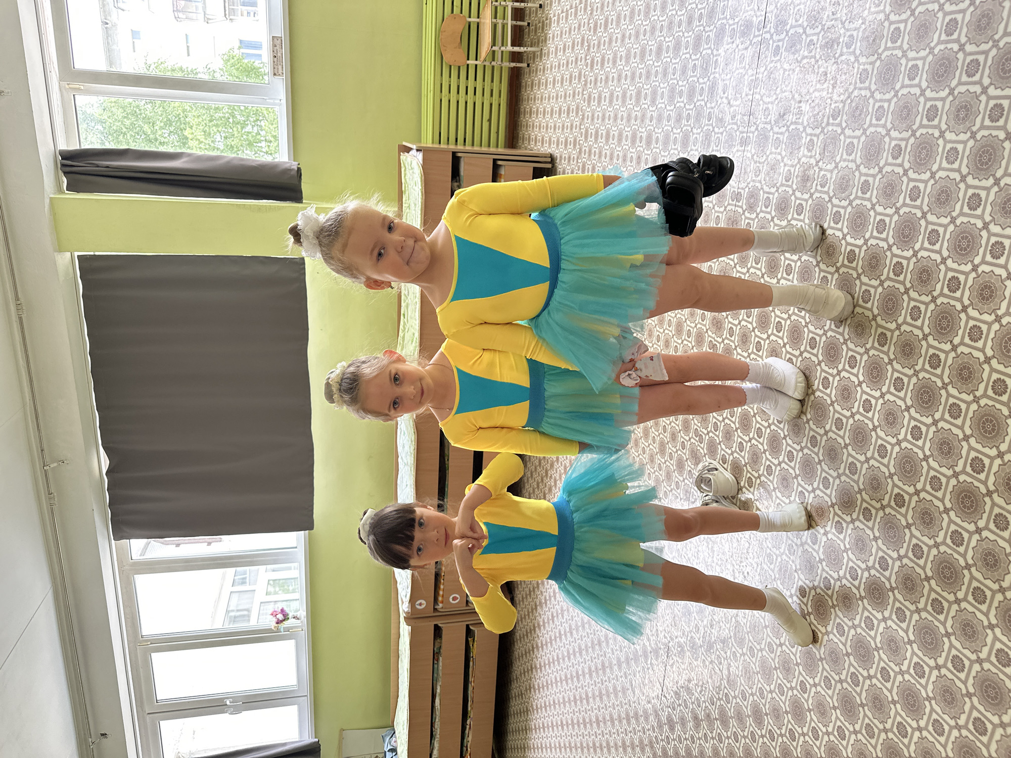 Three young girls are positioned in a vertical line, all facing the camera. They wear identical yellow and turquoise leotards with matching turquoise tulle skirts and white socks. The top girl has blonde hair styled in a high bun and carries black ballet shoes. The middle girl also has blonde hair in a bun. The bottom girl has dark hair with bangs. They are indoors, standing on a tiled floor with a geometric pattern. In the background, there is a window with gray curtains, a green wall, and wooden furniture, indicating the location is likely a dance studio or classroom in a building. The scene appears to be in Chaikovsky, Russia.