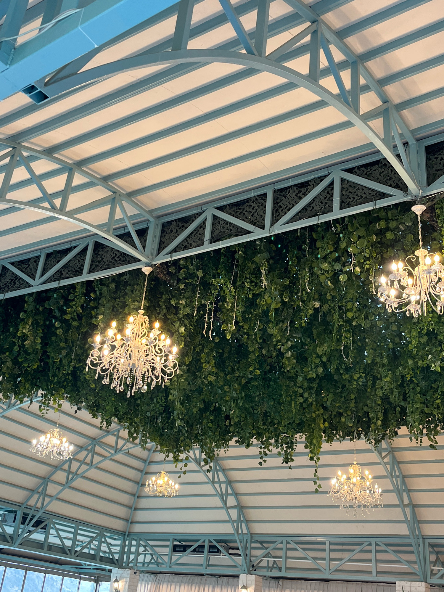 The image shows an interior scene with a light blue and white ceiling structure made of metal beams. Several ornate chandeliers are hanging from the ceiling. A thick curtain of green foliage, possibly artificial ivy, drapes down from a higher level of the ceiling, partially obscuring some of the chandeliers and the ceiling structure. The foliage creates a lush, naturalistic backdrop. The overall impression is one of an event space or restaurant decorated with a mix of elegant and natural elements. The scene is set in the city of Garni, Armenia, suggesting a venue or establishment within that location. No people are visible in the image.
