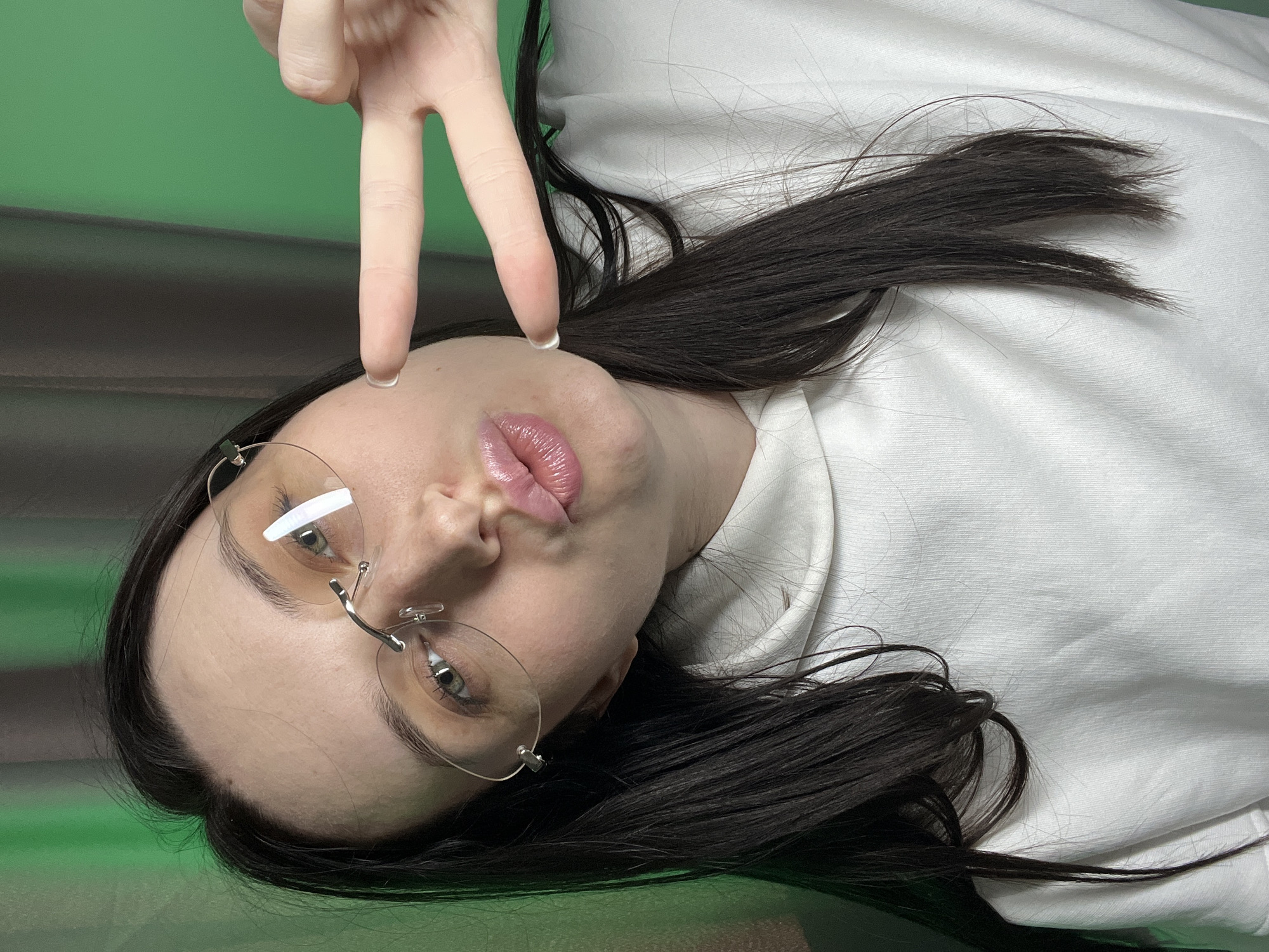 A close-up, chest-up portrait features a female subject with long, dark, straight hair, fair skin, and green eyes, looking directly at the camera. She wears rimless eyeglasses. Her lips are puckered in a kissing gesture. Her right hand is visible on the right side of the frame, making a 'peace' or 'victory' sign with the index and middle fingers extended. She is dressed in a white hooded sweatshirt. The background consists of a plain green wall to the right and a dark grey or brown curtain to the left, indicating an indoor setting. The specific location within the city of Tyumen, Russia, cannot be determined from the visual information provided.