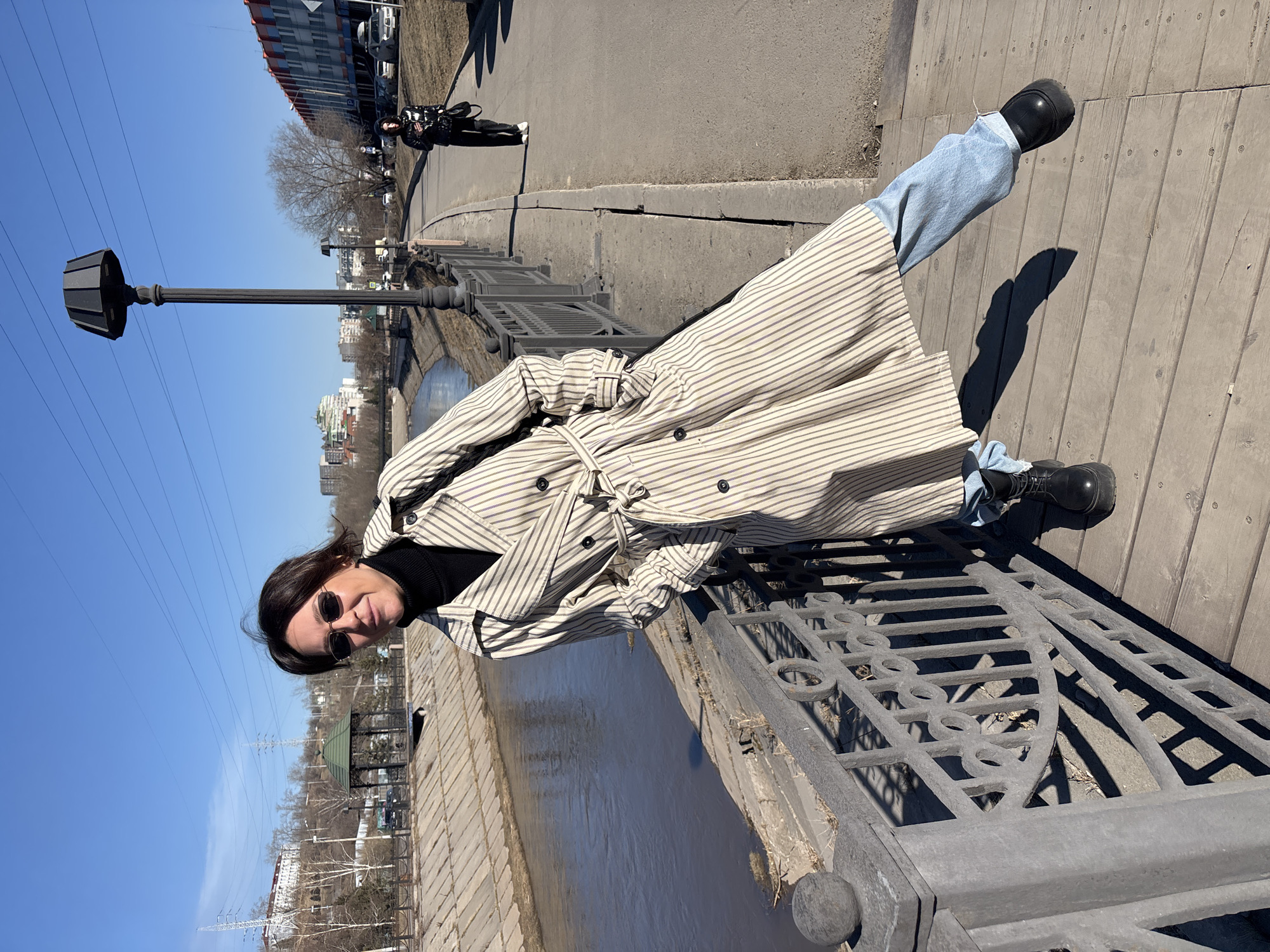 A woman stands on a bridge in Krasnoyarsk, Russia. She wears a striped trench coat, black turtleneck, blue jeans, and black boots. She also wears sunglasses. The bridge has a metal railing and wooden planks. A river flows below. In the background are buildings, trees, and a blue sky. A lamp post stands to the left of the woman. A person on a scooter is in the background.