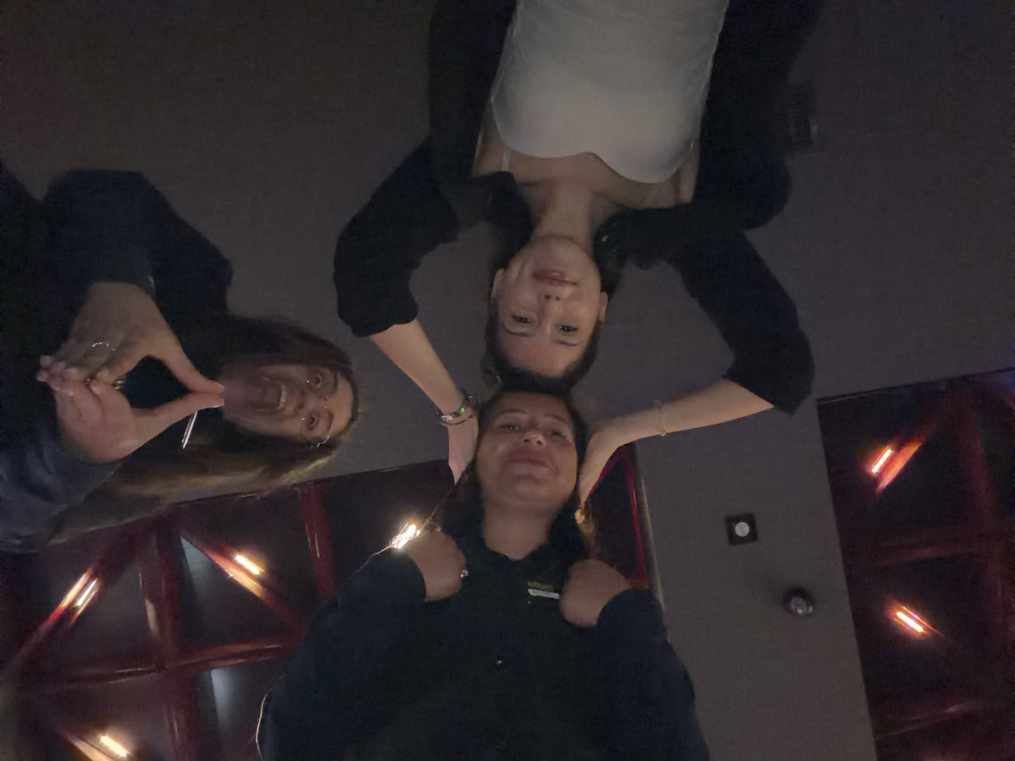 Three individuals are lying on their backs, with their heads in close proximity, forming a triangular arrangement. One person, in the foreground and slightly to the right, is wearing a white strapless top and a black open cardigan. Their head is turned to their left, and they are smiling. To their left, another individual, wearing a dark-colored top, is looking upwards with a neutral expression. The third person is at the top of the frame, with their head tilted downwards. They are wearing a dark top and have glasses. Their hands are clasped together above their head, forming a heart shape. The background appears to be a dark, interior space with some architectural elements visible, including what looks like a lattice-like structure with lights behind it. There are also lights integrated into the surface below the individuals, which is dark and reflective.
