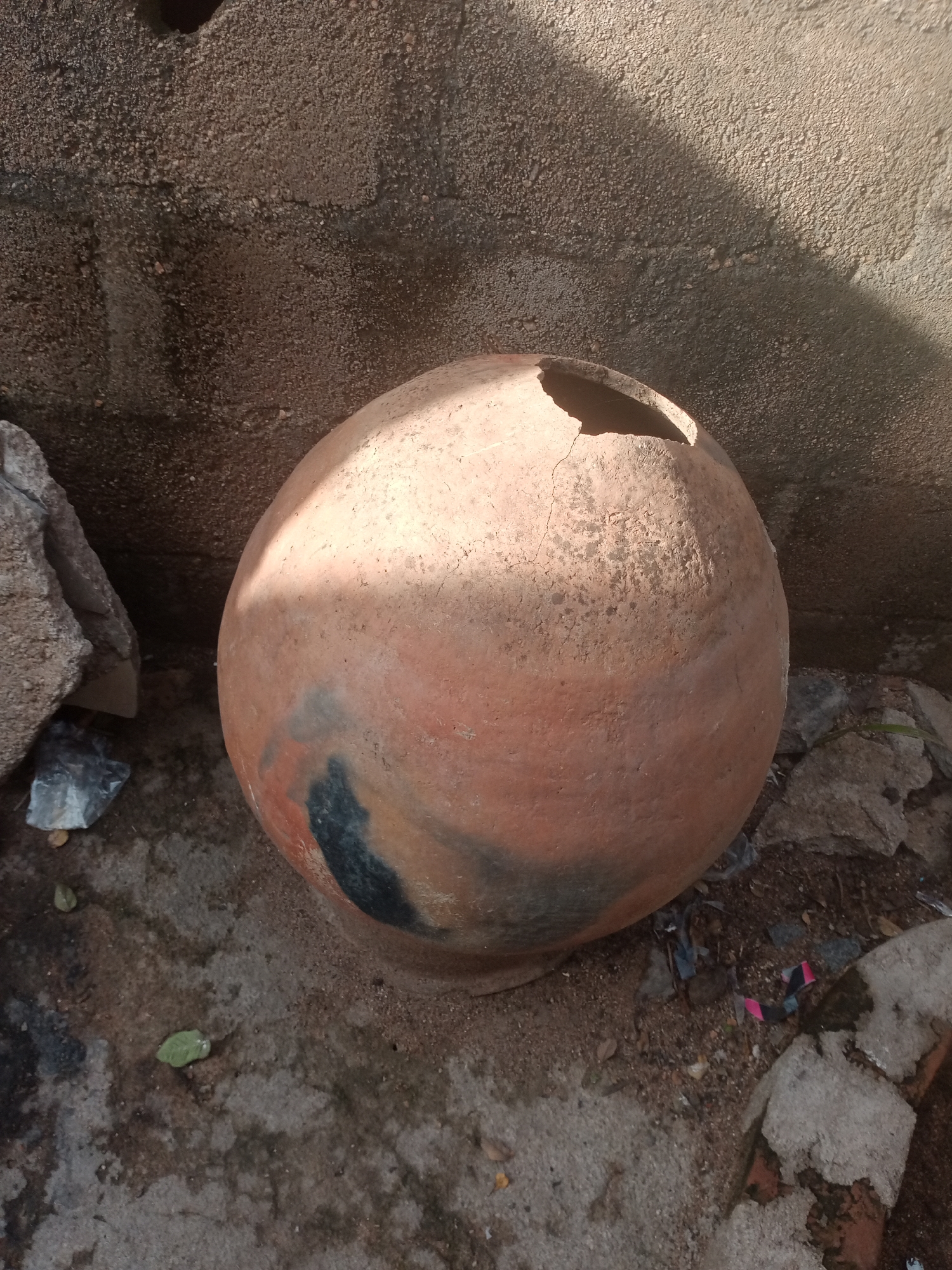 The image shows a large, round, terracotta pot with a damaged rim, sitting on the ground next to a rough, concrete wall. The pot has a mottled appearance with areas of darker color, possibly due to weathering or staining. Debris, including rocks and scattered litter, is visible on the ground around the pot. Sunlight casts a shadow on the wall and a portion of the pot. The setting appears to be an outdoor area, likely in a less-maintained part of Jalingo, Nigeria, given the texture of the wall and the general appearance of the environment. There are no people or animals present in the image.