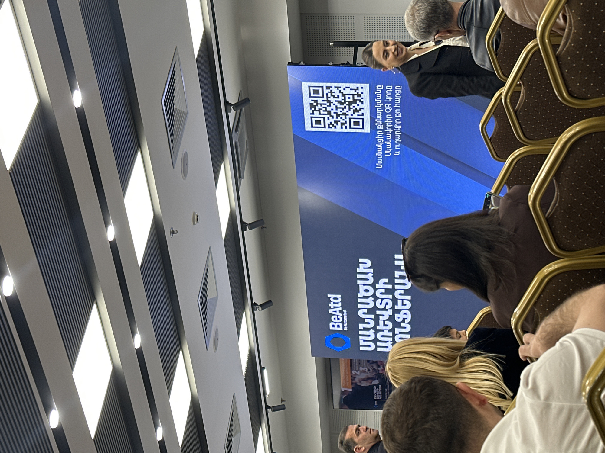 The image shows a gathering of people, possibly at a conference or seminar in Yerevan, Armenia. A projection screen displays a QR code and the text "ՄԱՆՐԱԾԱԽ ԱՌԵՎՏՐԻ ՈՆՖԵՐԱՆ...", along with the brand "BeAtd, Be Automated." Some individuals are seated in chairs, facing the screen. A woman can be seen smiling and looking towards the audience. The ceiling features a pattern of lights and ventilation systems.