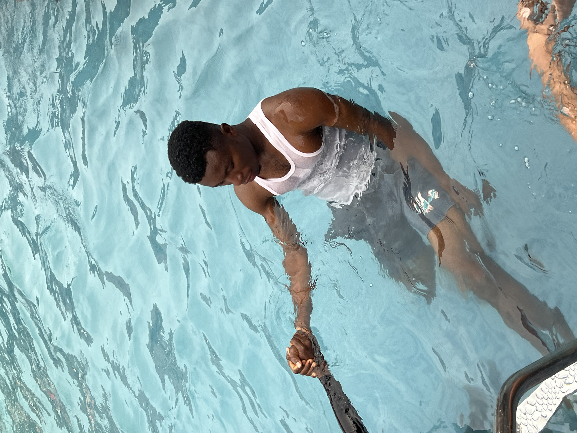 The media file captures an overhead view of a young man in a swimming pool, partially submerged in clear, light blue water. He appears to be floating or gently moving, wearing a white sleeveless top and dark shorts. His dark, short curly hair is visible, and his face is turned downwards towards his right hand. This hand is clasped firmly by another person's hand, which extends into the frame from the lower left, suggesting an act of support, guidance, or connection. Part of another person's legs and lower torso are visible near the upper right edge of the pool, indicating multiple people are present. The water exhibits strong ripples and reflections, suggesting a bright, sunny daytime environment. A metal handrail for pool entry/exit is visible in the bottom right corner. The activity appears to be swimming, floating, or a supervised water interaction. The setting is an outdoor swimming pool located in Nkpor, Nigeria. There is no visible text in the image.