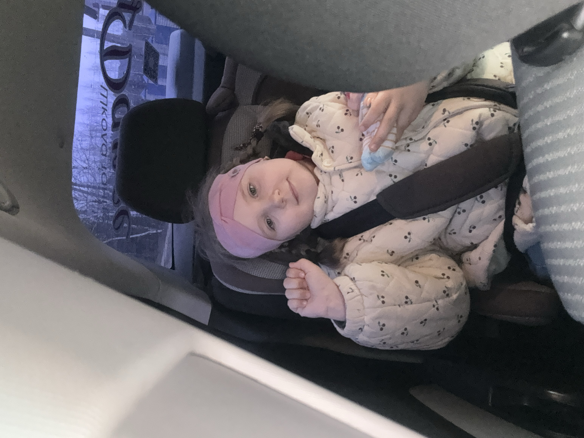 A young child is secured in a car seat in what appears to be the back of a vehicle. The child wears a light-colored quilted jacket with a small cherry pattern, a pink headband, and braided hair. The child is buckled in with the car seat's harness and holds a soft, light-blue and white striped object, possibly a toy or blanket. A window with some lettering, "Фоки, По", and the reflection of a building can be seen through the car window. This indicates a location in the city of Фоки, Russia. The overall environment suggests a car interior.