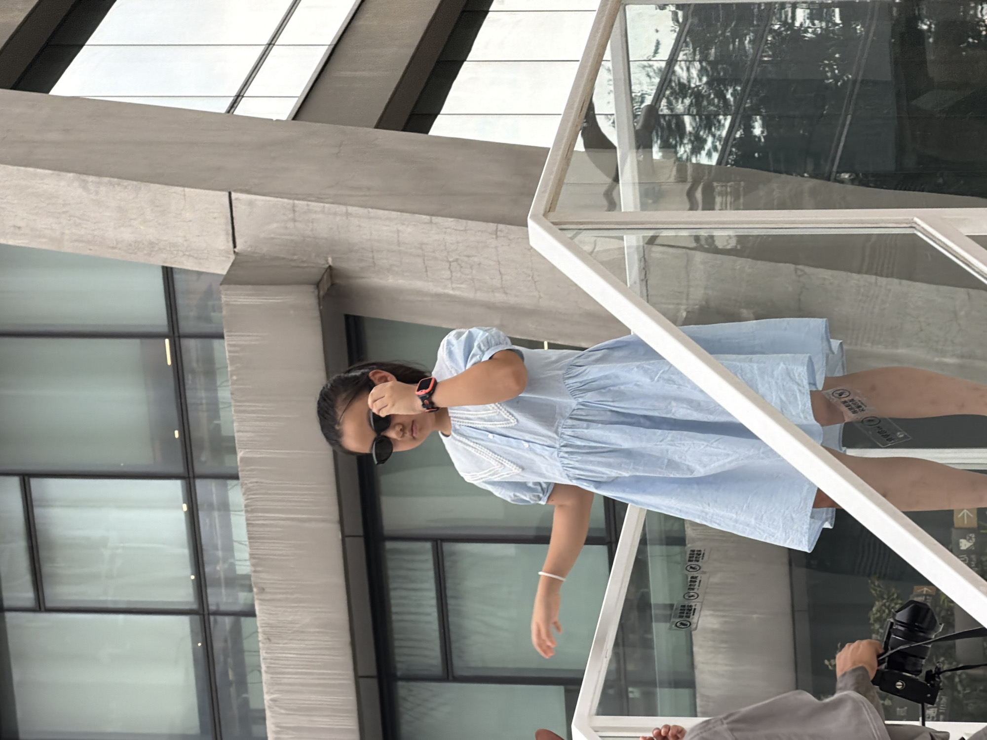 In Yuzhong District, China, a young East Asian girl wearing sunglasses and a light blue dress stands on a white staircase. She has her right hand near her face, adjusting her sunglasses, and a watch on her left wrist. The staircase has a glass railing. In the background, a modern building with large windows is visible, along with a concrete architectural element. A person holding a camera is partially visible on the left side of the image.