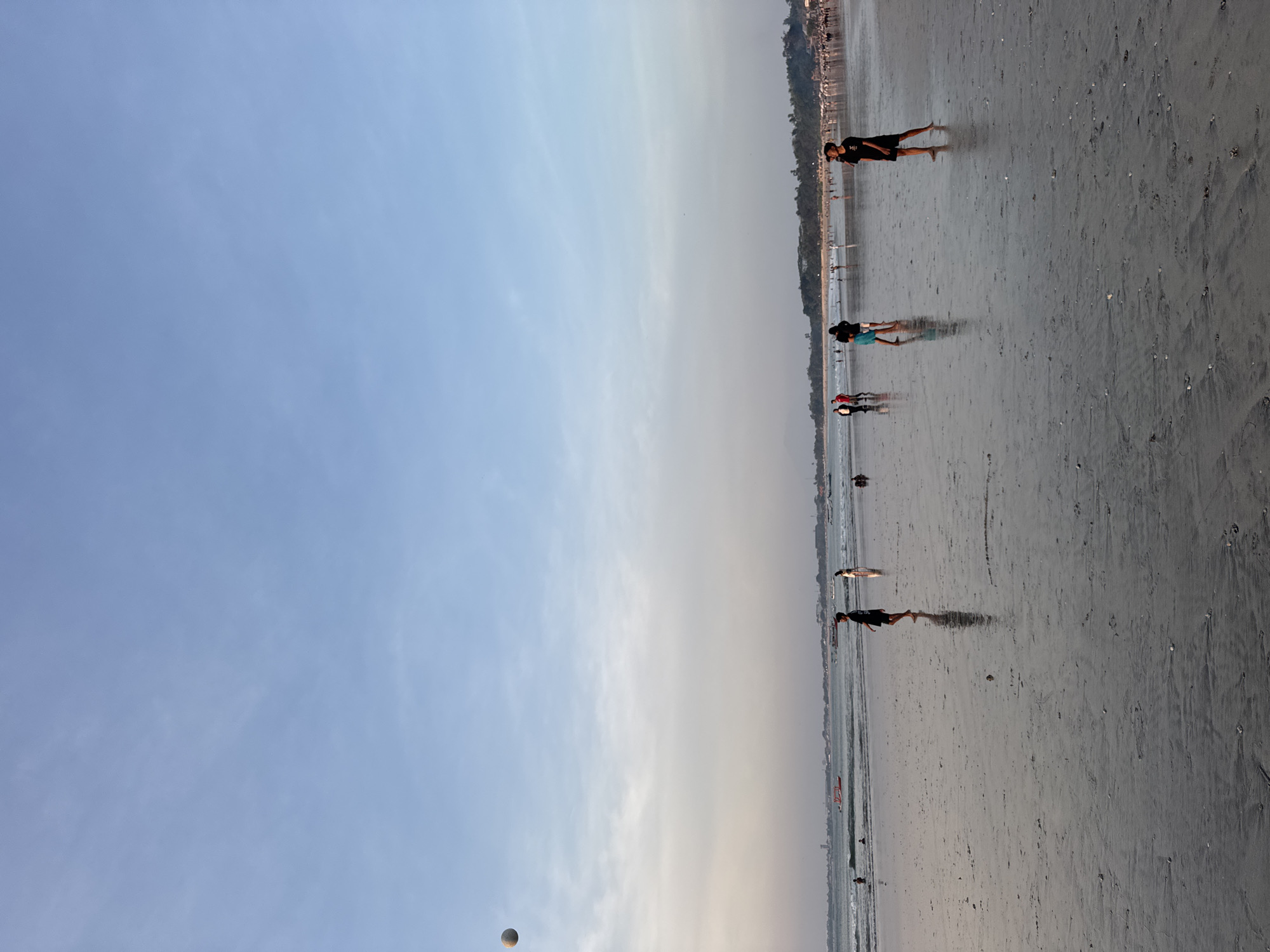 This is a medium shot, eye-level photograph of Kuta Beach in Kuta Selatan, Indonesia, taken at dusk. The sky is a pale blue with wispy white clouds. A round, pale object, possibly a ball, is suspended in the air in the upper left quadrant of the image. The wet sand on the beach reflects the sky and the figures present. In the foreground, a person in a black t-shirt and shorts stands facing left, their reflection visible on the wet sand. In the mid-ground, several other figures are scattered across the beach, some walking and some standing. To the left, there are small waves lapping at the shore, and further out, several boats are visible. The background shows a long stretch of beach lined with trees and buildings. The overall atmosphere is peaceful and serene, with a few people enjoying the beach at sunset.