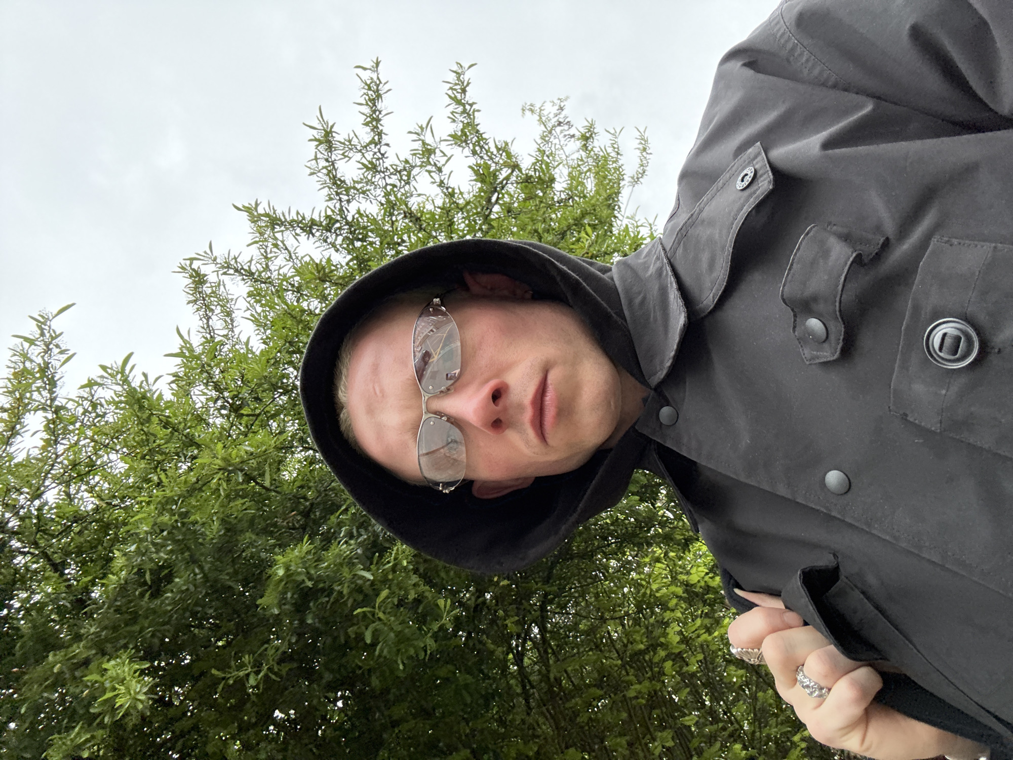 This low-angle selfie captures a fair-skinned individual outdoors against a backdrop of green trees and an overcast sky. The person, appearing to be male, is seen from the chest up, wearing a black hooded jacket or coat with visible flap pockets and dark buttons. Their short blonde hair is partially visible beneath the hood. They wear silver-framed glasses with highly reflective lenses that obscure their eyes, showing reflections of the surrounding trees and sky. Their lips are slightly parted, and their expression is neutral as they look directly into the camera.
The person's right hand is visible in the lower right foreground, clasped over a part of their jacket, adorned with several silver rings featuring textured designs on the fingers. The background is dominated by dense green foliage of trees, suggesting a park, garden, or natural area, consistent with green spaces found in London. The sky is a uniform light gray, indicating an overcast or cloudy day. No text is visible in the image.
