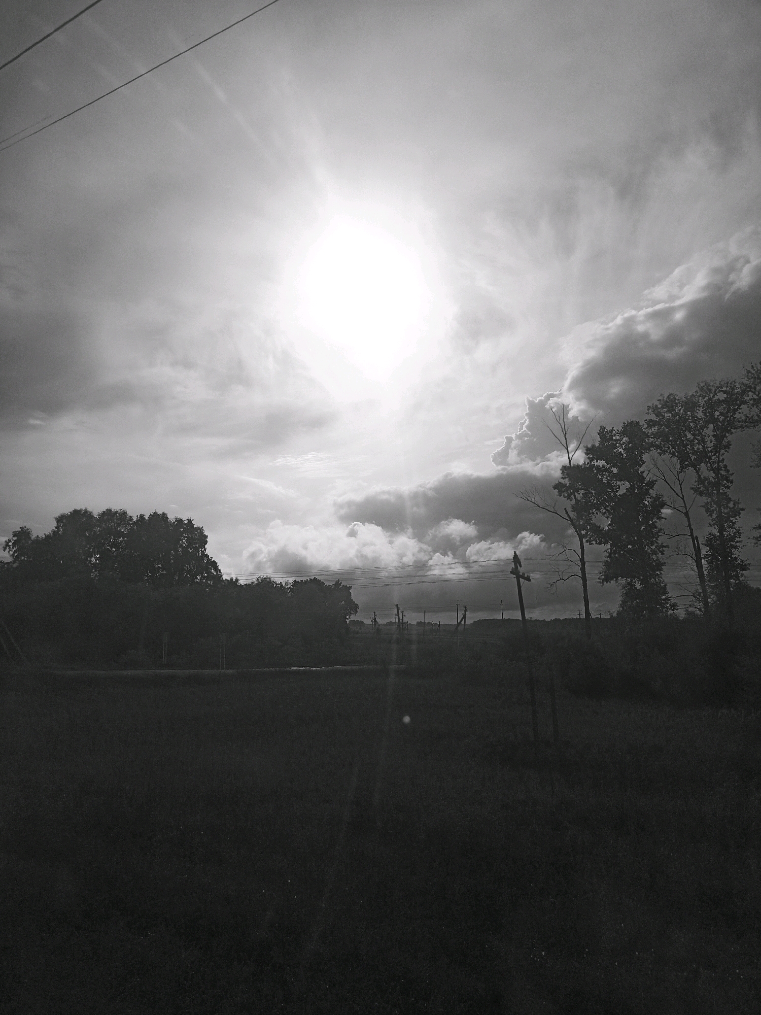 The image depicts a landscape scene at dusk, with the sun low in the sky, casting bright rays through a cloudy atmosphere. In the foreground, a dark, silhouetted field stretches across the bottom half of the frame. In the middle ground, a line of dark trees and vegetation runs horizontally, obscuring the horizon. Several utility poles and wires are visible, some with a bird perched on one of them. The background shows a hilly terrain under a dramatic sky filled with clouds and the setting sun. The overall impression is a rural or semi-rural environment under a moody sky. There is no indication of the location being in the city of Kurundus, Russia; the image does not provide any specific geographical markers to confirm or deny such a location.