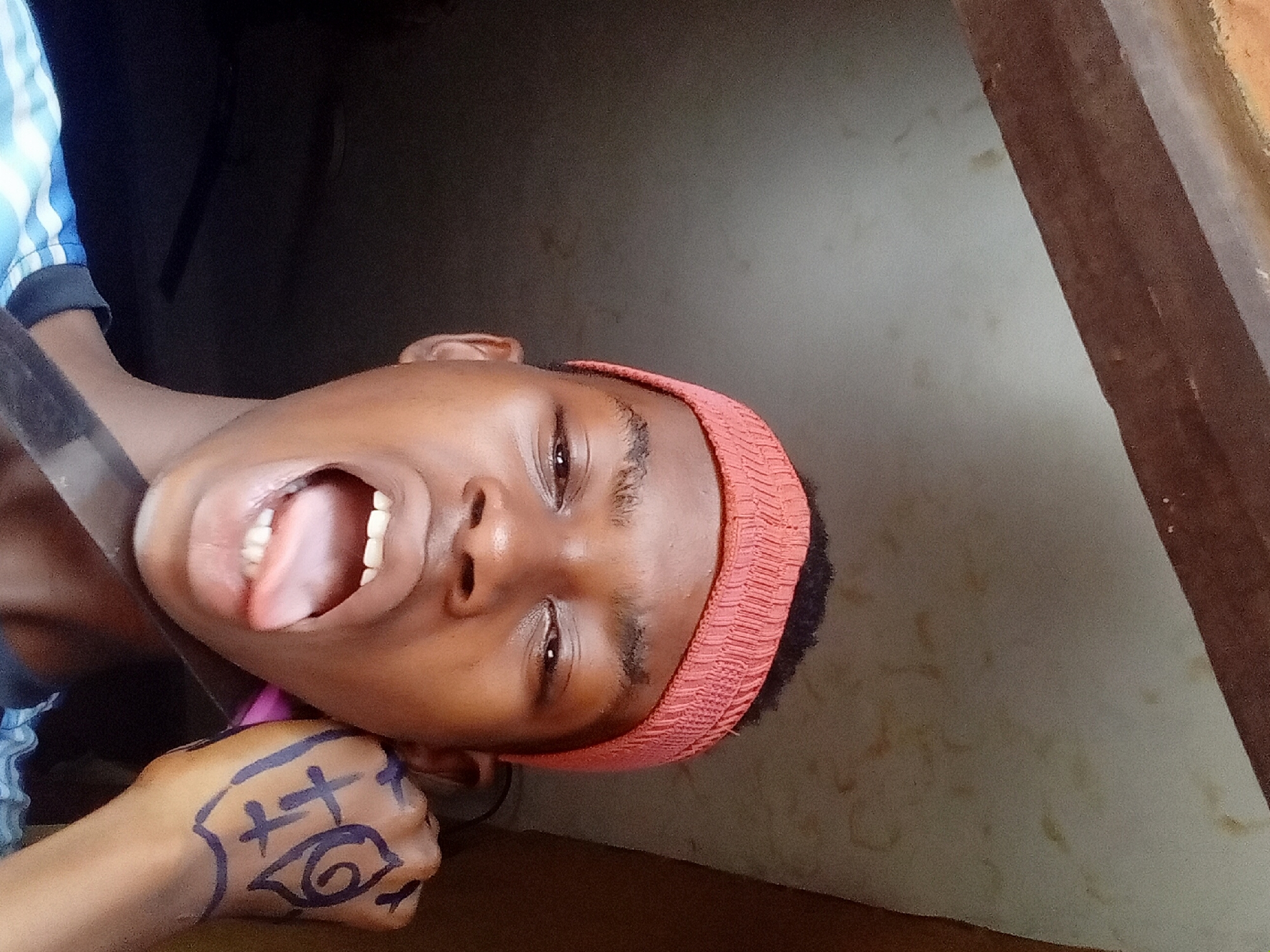 A person with dark skin, wearing a reddish-orange headband and a blue and white striped shirt, is positioned in what appears to be a doorway. The person's mouth is open, tongue extended. Their fist, marked with black ink, is raised. The background includes a dark, interior space and an adjacent wall. This scene is likely located in the city of Onitsha, Nigeria.