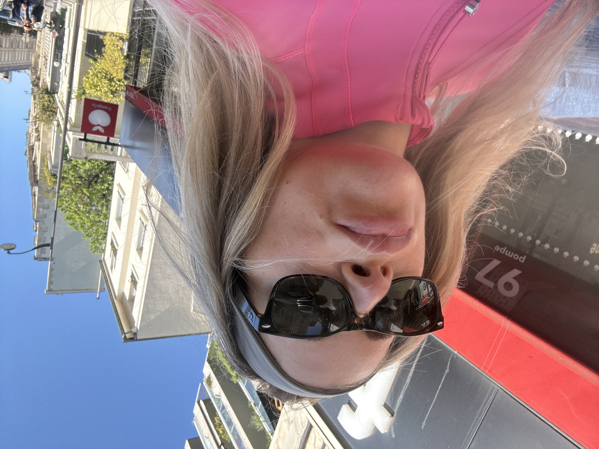 This image is a close-up, upside-down selfie of a person, likely taken outdoors on a bright, sunny day in an urban setting.
The subject has blonde hair, fair skin, and is wearing dark sunglasses with "Ray-Ban" branding visible on the frame. A light gray headband is worn beneath the sunglasses. They are dressed in a vibrant, hot pink athletic top or jacket, featuring visible stitching and a metallic zipper pull. Their lips are slightly parted.
The background, largely inverted due to the image orientation, reveals an urban landscape. Multi-story buildings with light-colored facades and windows are visible, some adorned with balconies. Green foliage from trees and bushes suggests a pleasant, leafy environment. The sky is a clear, brilliant blue, indicating excellent weather.
Key textual and contextual elements confirm the location as Paris, France. A red and white sign for "Franprix," a common French supermarket chain, is clearly visible. Additionally, a partial red and black sign shows the word "pompe" (French for "pump") and the number "97" (both appearing upside down), suggesting the presence of a gas station or possibly a public service like a bike or scooter rental station. A street light fixture is also visible against the clear sky.
The overall impression is a casual, sunny snapshot from a city outing.