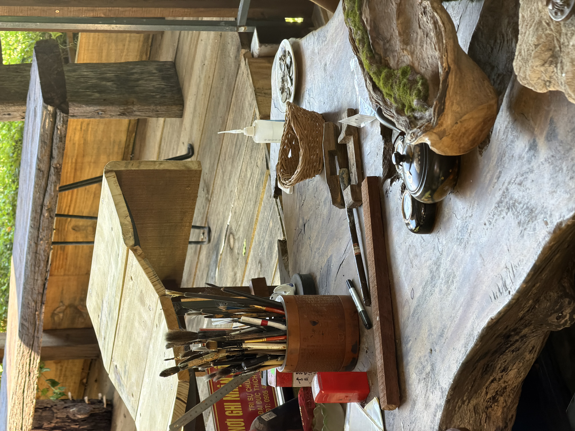 This is an indoor scene set in what appears to be a workshop or art studio. The focal point is a wooden tabletop covered with various art supplies and decorative objects.
Objects on the table include a cylindrical container filled with paintbrushes, a ruler, a red container, and other miscellaneous items. There's also a wooden box-like structure, possibly a small easel or storage unit. Toward the right, a unique carved wooden piece with moss growing on it sits beside a decorative teapot.
The background consists of wooden structures, suggesting the interior of a rustic building. Light filters in from the left side of the frame, partially illuminating the scene.
An OCR snippet on one of the objects includes Vietnamese text with phone number, suggesting the scene could be somewhere in Vietnam, likely Đà Nẵng.