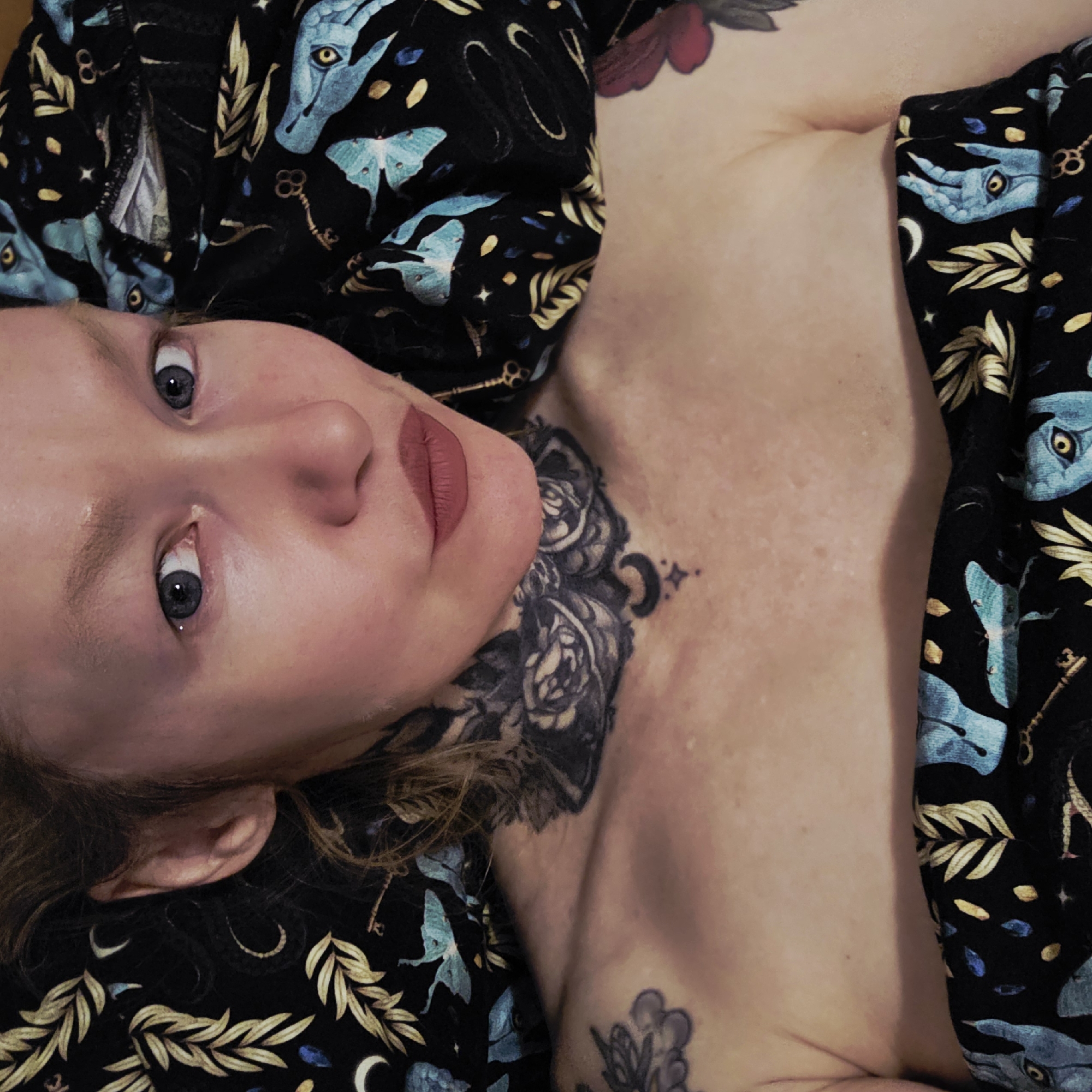 A woman with fair skin and blue eyes lies on a black fabric adorned with blue moths, golden leaves, keys, crescent moons, and blue animal heads. She has a large floral tattoo on her neck and smaller tattoos on her chest. Her lips are painted a matte red. The background is out of focus. The location is not discernible from the image.