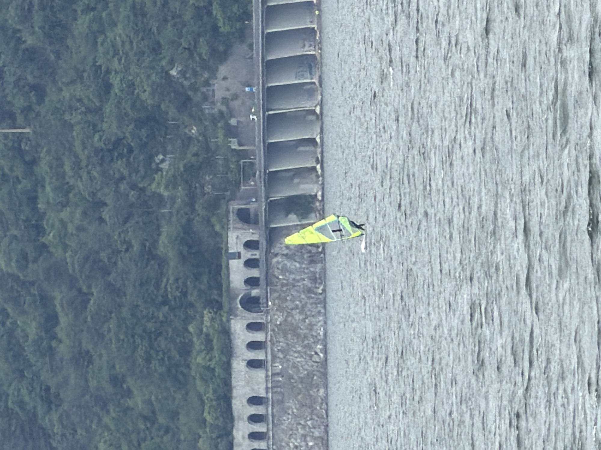 This vertically oriented image presents an aerial or elevated view of a waterside scene in Valmadrera, Italy, featuring a combination of natural landscape and man-made structures.
The right side of the frame is dominated by a large body of water, likely a lake, characterized by a rippled, greyish surface, suggesting the presence of wind. On this water, a prominent bright yellow and green triangular object, resembling a sail, is visible. This object is likely part of a windsurfer or a small sailing craft, indicating water sports activity. A person on the craft is not clearly discernible.
The left side of the image features a dense, lush green forested hillside or mountain slope, providing a natural, verdant backdrop.
Separating the water from the forested land is a substantial man-made structure along the bank. This structure appears to have two distinct sections. Towards the upper part of the bank, there is a segment composed of several large, rectangular, open-topped basins or compartments, possibly part of a dock, industrial facility, or a specialized retaining wall. Further along the bank, closer to the water, a more elaborate stone or concrete structure with a series of uniform arches is visible, resembling an old bridge, aqueduct, or a fortified waterfront. Small, indistinct figures or objects can be seen on the bank near these structures.
The scene is captured during bright daylight hours, under clear weather conditions. The overall impression is one of a scenic location where nature and human construction coexist, facilitating outdoor recreational activities. No visible text is present in the image.