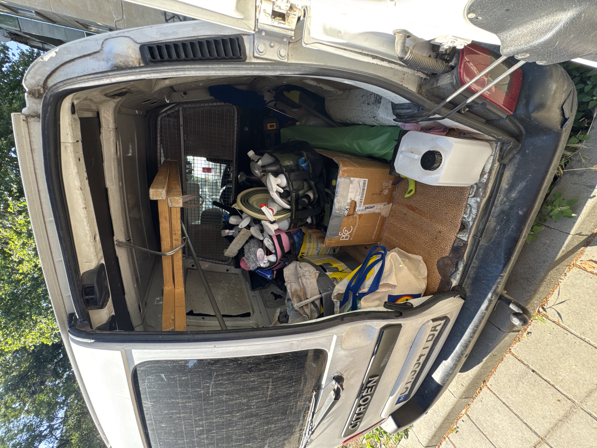 The image shows the open rear of a white commercial van, likely a Citroën, parked on a paved street in Plovdiv, Bulgaria. The van's interior is cluttered with various items, including cleaning supplies, bottles, bags, and what appear to be some cleaning tools like brushes and a dust mop. A metal cage is visible separating the cargo area from the cabin. The van is parked next to a sidewalk with grass growing through cracks. The day appears sunny, with trees visible in the background. The van's license plate is partially visible and appears to have Bulgarian registration.