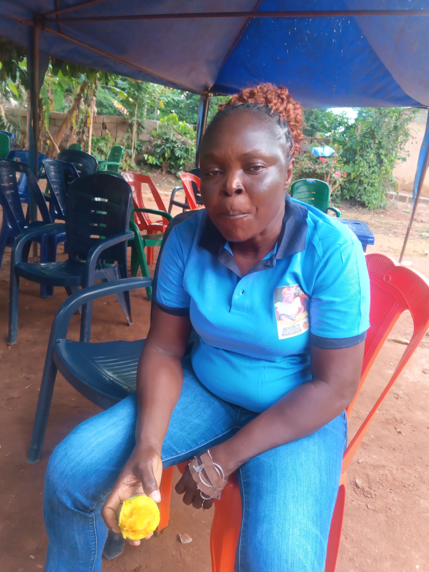 A dark-skinned woman with reddish-brown, curly hair pulled back is seated on a dark grey plastic chair. She is wearing a light blue polo shirt with a black collar and cuffs, and blue denim jeans. Her expression is neutral, with her mouth slightly open. In her right hand, she holds a partially eaten, bright yellow fruit, likely a mango. On her left wrist, she has several light-colored bands.
A patch or badge is visible on the upper left side of her polo shirt, featuring a picture of a woman and text that reads "SENATE BENADETH CHINWENDU MADUAKOR."
The scene is outdoors, beneath a blue tarp or canopy, suggesting a temporary shelter. Numerous other plastic chairs in shades of blue, red, green, and black are scattered in the background, some empty. The ground is reddish-brown dirt. Behind the chairs, lush green vegetation, including what appears to be banana trees, and a concrete block wall are visible. The lighting suggests it is daytime. The overall setting appears to be a casual gathering or waiting area in a warm, outdoor environment consistent with Eziudo, Nigeria.