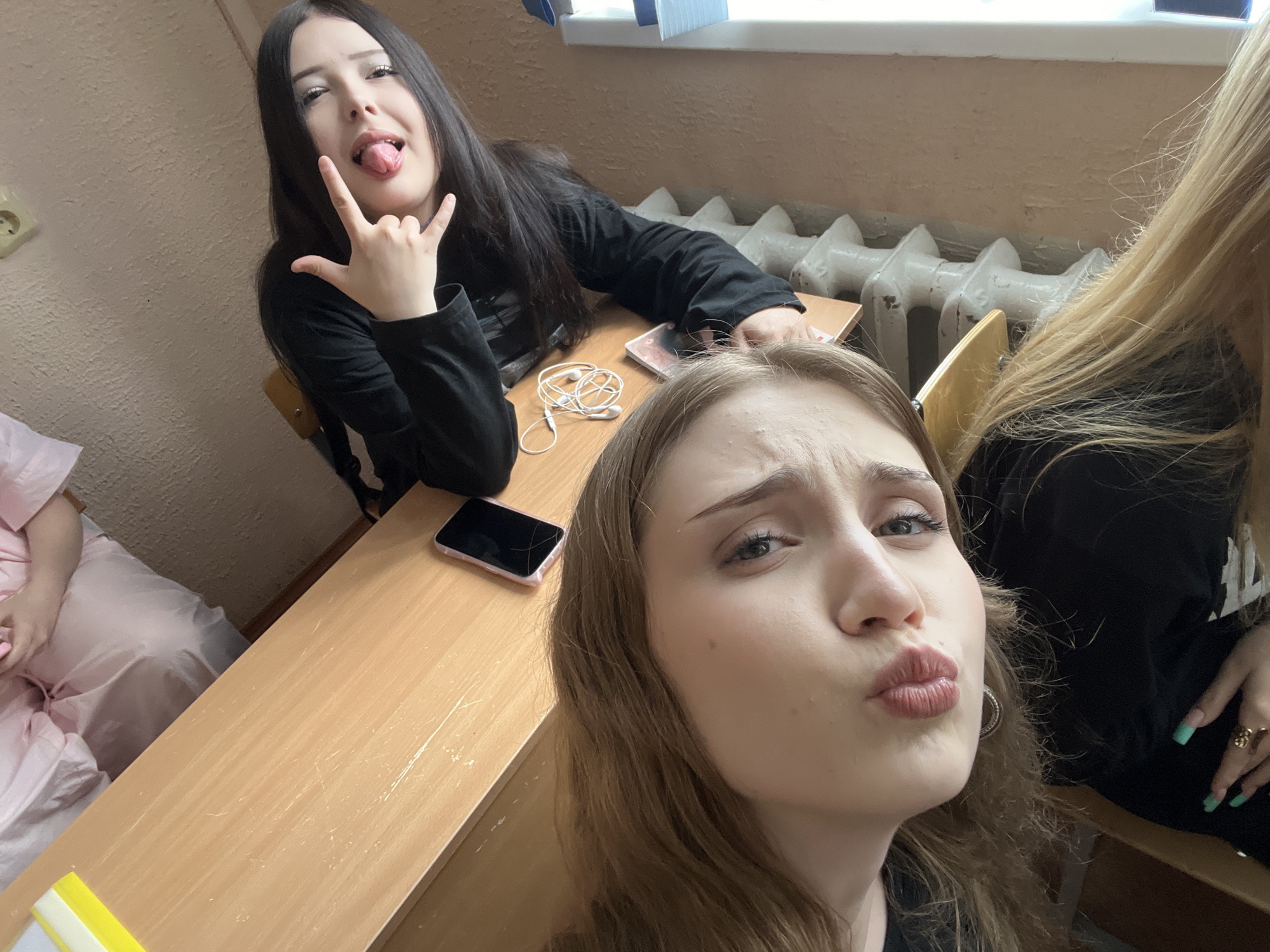 The image depicts a selfie taken by two young women in a classroom environment. The woman in the foreground, with light brown hair, is puckering her lips and looking towards the camera. Behind her, a woman with long dark hair is sticking out her tongue and making a rock-on hand gesture. They are seated at wooden desks, with a white radiator visible in the background. A mobile phone and a pair of white earbuds are on the desk. A person in a pink garment is partially visible on the left side of the frame. The overall scene suggests a casual and playful moment captured within an educational setting. The location is identified as a classroom in Sterlitamak, Russia.
