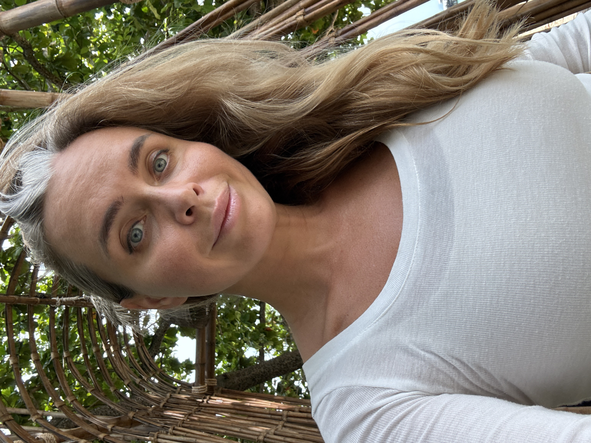 In Ko Samui District, Thailand, a woman with blonde hair and blue eyes is captured in a close-up selfie. She is wearing a white long-sleeved shirt. The background consists of lush green foliage and a woven bamboo structure. The lighting suggests it is daytime.