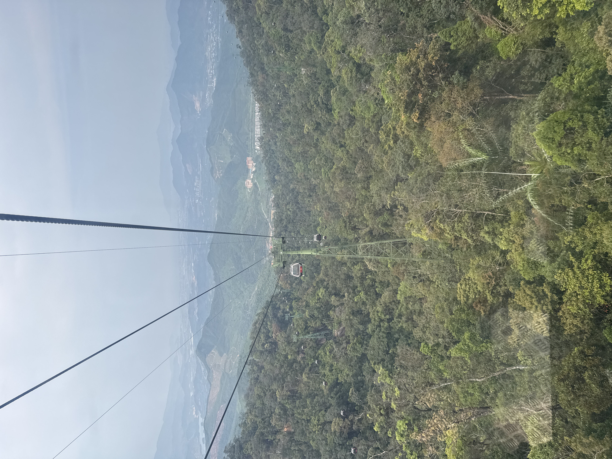 The image captures a panoramic view from a gondola lift, looking down over a vast, forested mountain landscape. The perspective is from inside a gondola cabin, with the dark support cables of the lift system prominent in the foreground, stretching diagonally across the frame.
In the midground, the intricate gondola lift infrastructure is visible, featuring sturdy green metal towers (pylons) supporting the cables. Multiple gondola cabins, including one distinct red and white cabin, are seen suspended along the lines, either moving towards or away from the nearest tower. Other smaller, grey cabins are scattered further along the route, highlighting the scale of the lift system.
Below the gondolas, dense, lush green forest covers the mountain slopes, indicating rich vegetation. The varied shades of green suggest a diverse tree canopy. Further in the distance, a valley floor with scattered buildings and structures can be faintly discerned. Beyond the immediate mountains, a series of hazy, blue-grey mountains extend into the background under a light, uniform sky, suggesting a vast mountainous region. The atmospheric conditions appear hazy or slightly overcast, softening the distant views.
The scene depicts a daytime view, showcasing the journey over a natural, rugged terrain. There is no discernible text visible in the image. Given the context, the image captures a scenic experience in a mountainous area, likely near Da Nang, Vietnam.