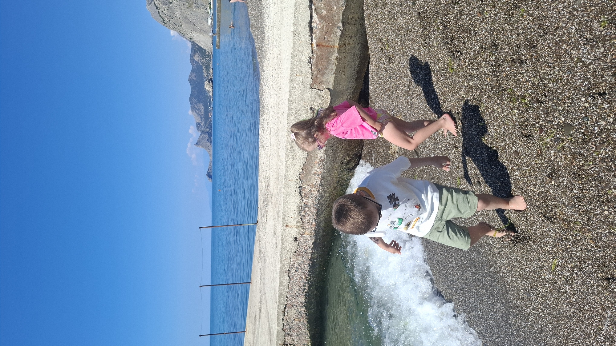 Two young children, a boy and a girl, are positioned on a pebbly beach next to a concrete structure on a bright, sunny day. The boy, with short light brown hair and wearing a white t-shirt with a graphic design of mountains and trees, along with olive green shorts, is bending down at the water's edge. He appears to be interacting with a small wave breaking on the shore. He has sunglasses perched on his head. The girl, with long light brown hair and dressed in a pink top over patterned shorts, stands slightly further back on the concrete structure, looking downwards.
The scene is set at a shoreline with clear blue water stretching to the left, where a distant rocky coastline or mountain range is visible under a cloudless blue sky. A weathered concrete structure runs parallel to the water, separating the sea from the coarse, pebbly beach. The bright sun casts distinct, elongated shadows of the children and the concrete, indicating either morning or late afternoon. Both children are barefoot. The white t-shirt worn by the boy has a graphic design that includes some faint, indecipherable text or symbols within the landscape.