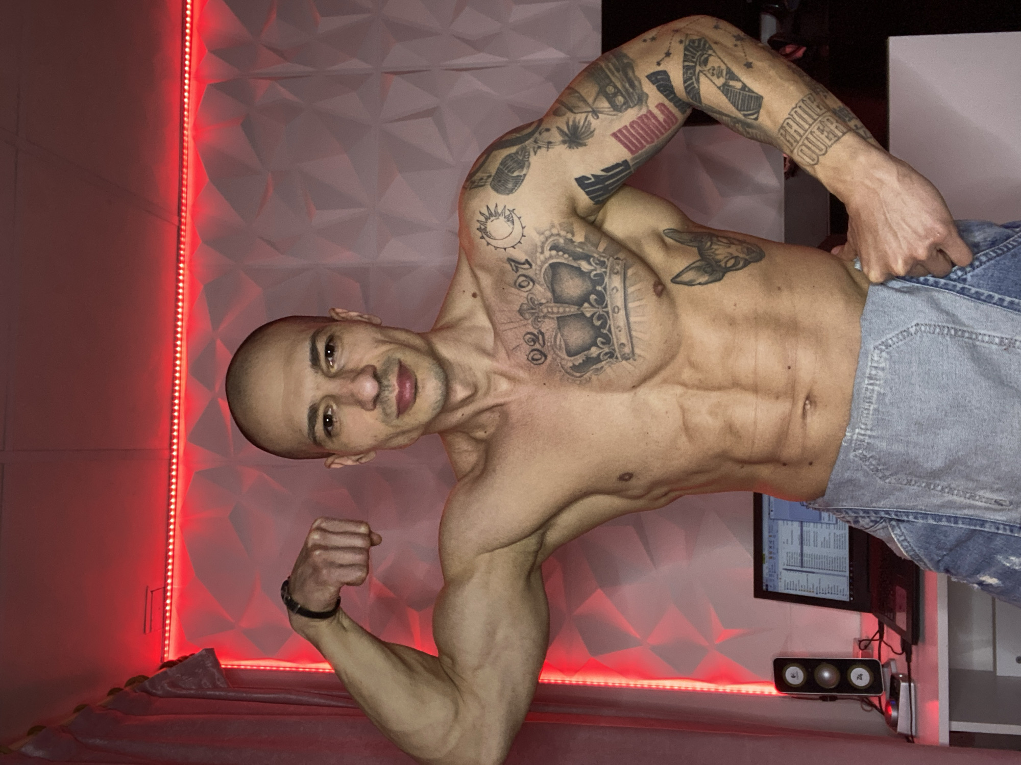 The image captures a bare-chested adult male standing in what appears to be a residential room in Pleven, Bulgaria. The individual, who is bald and possesses a muscular build with visible abdominal muscles, is positioned centrally, facing the camera directly. He flexes his left bicep, holding his left hand in a fist, while his right hand rests on his hip. He is partially dressed in light blue denim overalls, which are pulled down to his waist.
Distinct tattoos cover various parts of his upper body: a large crown design with the numerals "02 07" above it is on his left chest. His left shoulder and upper arm bear tattoos of a sun and moon, a microphone, and other abstract symbols. A cat's head tattoo is visible on his left side torso. His right arm features a full sleeve of tattoos, including a pharaoh's head, palm trees, a vintage-style vehicle, and the text "GAME OVER." A black wristband or watch is on his left wrist.
The background consists of a white wall adorned with geometric, possibly 3D, paneling. A horizontal strip of red LED lighting runs along the top of this wall, casting a red ambient glow throughout the visible area. To the left, a light-colored curtain or fabric hangs. A white desk is present on the lower left, supporting an open black laptop displaying data on its screen, a pair of black computer speakers with multiple drivers, and a small silver and black electronic device.