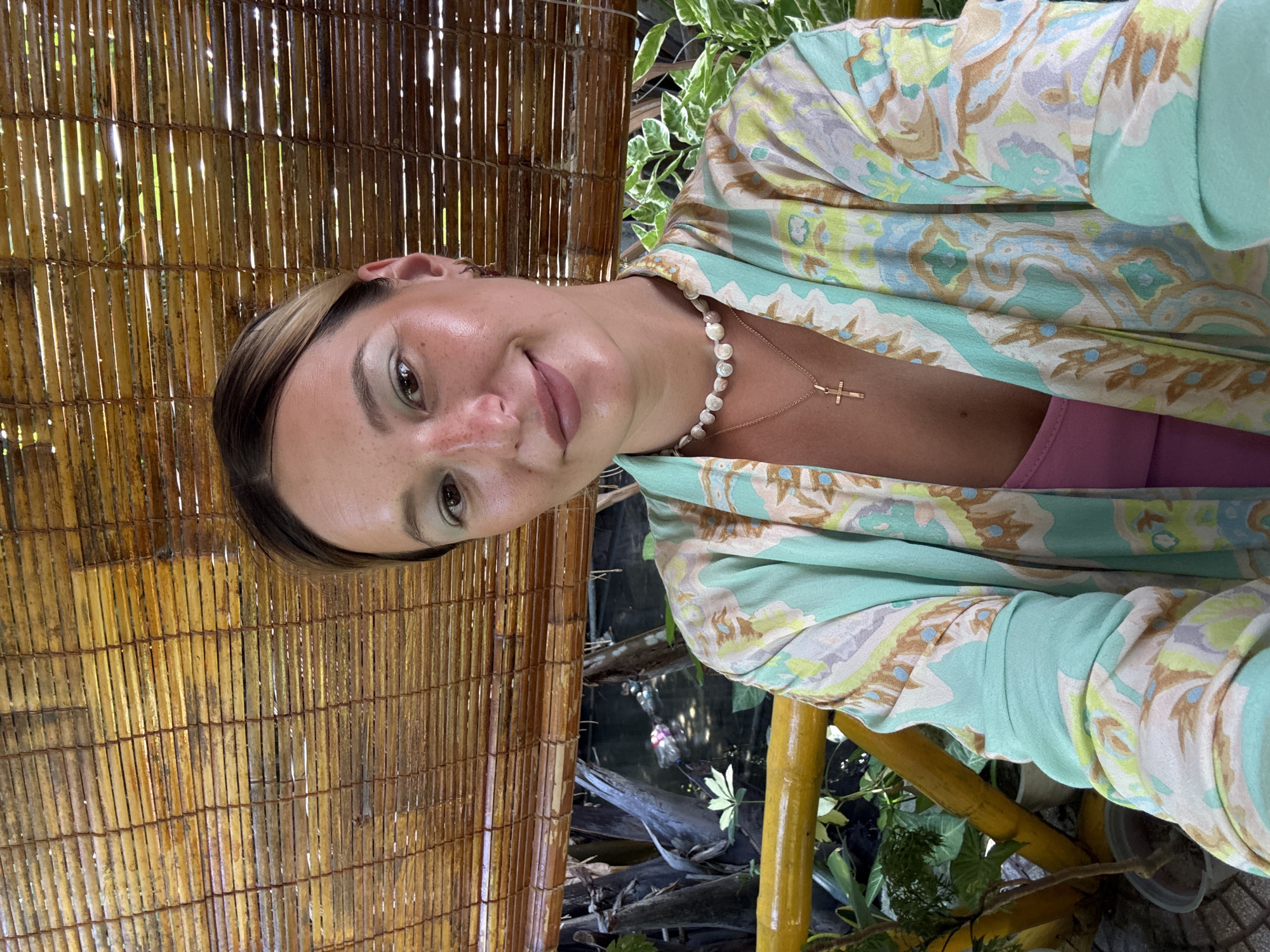 A young Caucasian woman with blonde hair pulled back and freckles is taking a selfie. She is wearing a mint green and cream patterned kimono over a pink top, a pearl necklace, and a gold cross necklace. She is smiling at the camera. The background consists of a bamboo blind and lush green foliage. The woman is seated outdoors on what appears to be a balcony or patio, with a yellow railing in the foreground. The scene suggests a tropical or subtropical environment, possibly a resort or a residential area with a view of nature. The specific location in Da Nang, Vietnam, is not explicitly identifiable from the visual elements alone.