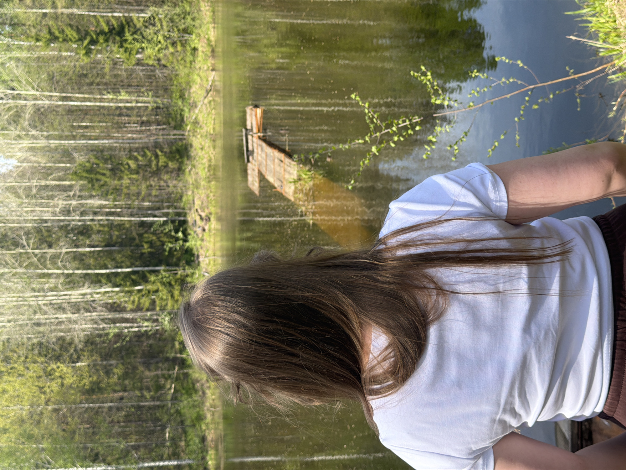 A person with long brown hair, wearing a white t-shirt, stands on a bank overlooking a body of water. The water reflects the surrounding trees and sky. A small wooden structure extends into the water from the opposite shore. A dense forest of slender trees stands along the left side of the image. The scene is set in Ural'skoe, Russia.