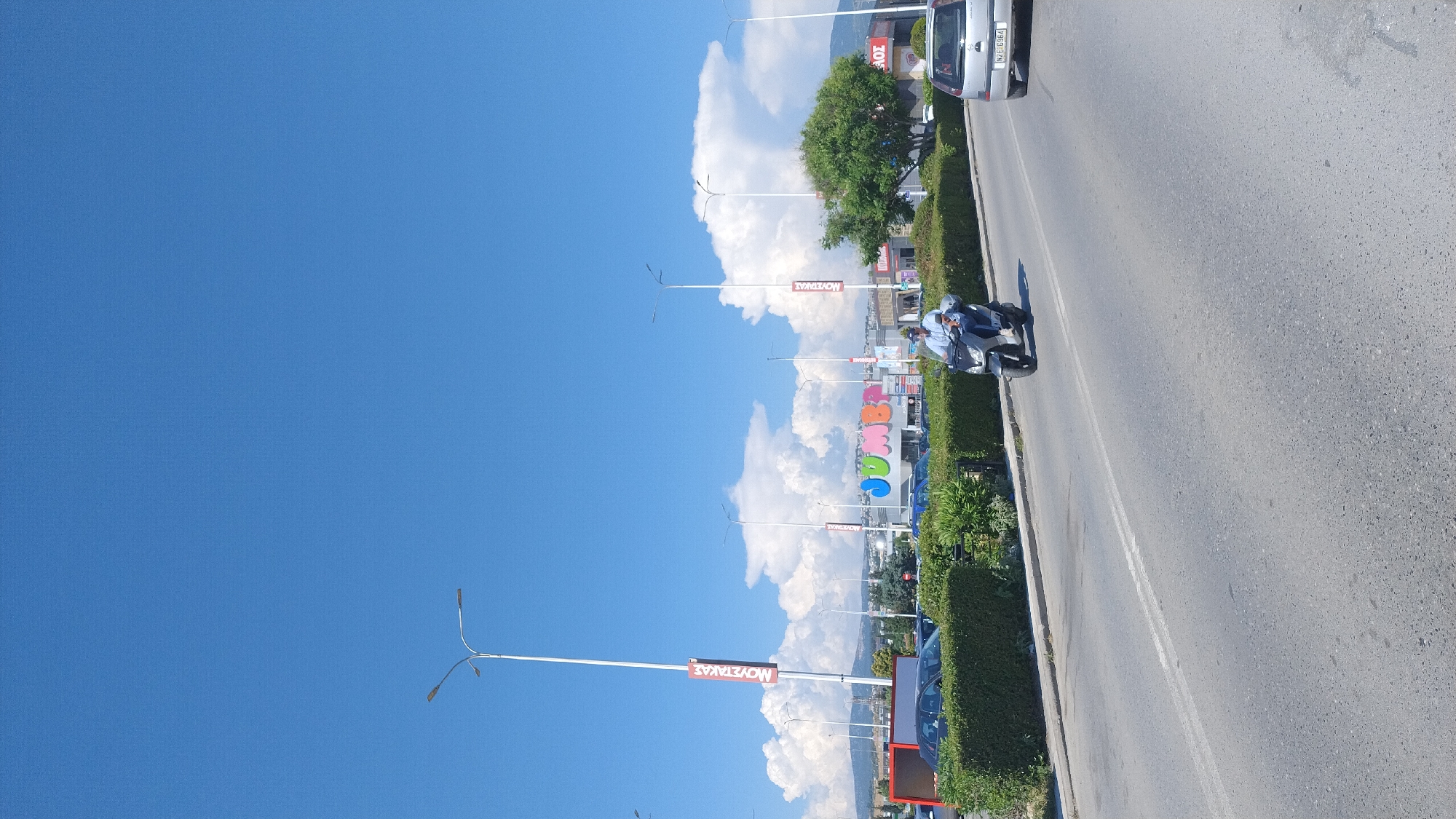 This image, taken in Pylaia, Greece, shows a street scene on a clear, sunny day. The sky is a vibrant blue, dotted with fluffy white clouds. On the right side of the frame, a paved road runs alongside a neatly trimmed green hedge. Cars are parked along the roadside, and a silver car with license plate "NKE-5964" is visible. A man on a scooter, wearing a light blue shirt, rides on the road.
In the background, several tall lampposts stand against the sky. Further back, large, colorful signs are visible, one of which spells out "jumbo" in large, individual letters in green, blue, and red. Other signs with red lettering read "MOVETAKAE," which appears to be Greek text. A few trees, including a large one with green foliage, add to the scenery. The overall impression is a bright and active commercial area on a pleasant day. The orientation of the image is sideways.