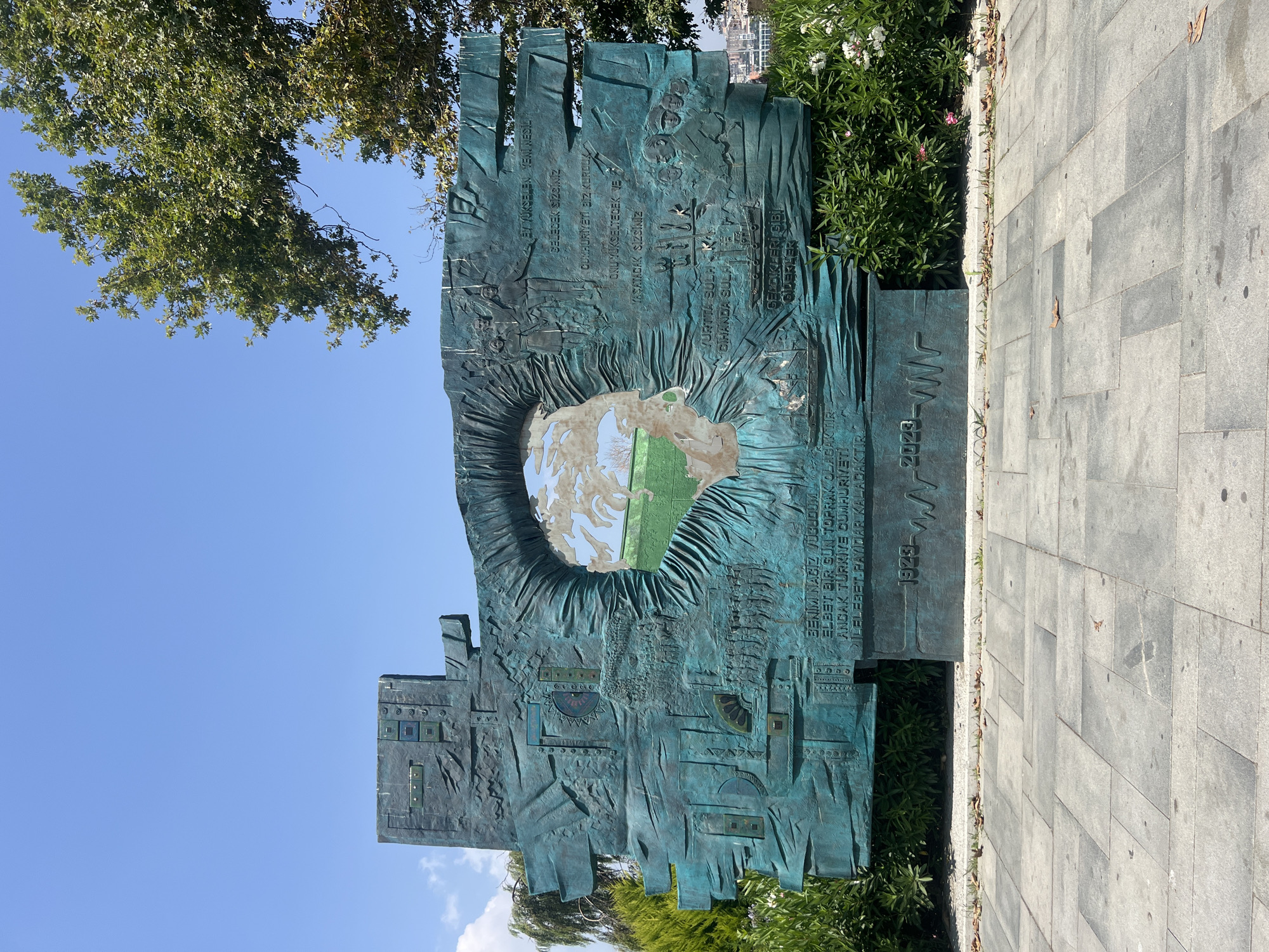 The image shows a large, teal-colored metal sculpture situated outdoors on a paved plaza. The sculpture is abstract and features a large, circular opening in the center that reveals a green field with a blue sky and clouds. The surface of the sculpture is textured and embossed with various images and inscriptions. To the left of the opening, there are depictions of gears, what appear to be circuit boards, and a procession of figures. To the right, there are more figures, including portraits of men, a ship, and inscriptions in Turkish. At the base of the sculpture, the years "1923" and "2023" are visible, separated by a waveform graphic.
The sculpture is surrounded by dense green foliage and bushes, with some white and pink flowers visible on the right side. A large tree with green leaves partially overhangs the sculpture from the upper right corner of the frame. The sky is a clear, bright blue with a few wispy clouds. The plaza is made of large, grey rectangular paving stones. In the background, beyond the sculpture and foliage, a cityscape with buildings is visible. This sculpture is located in Istanbul, Turkey, and is known as the "Republic of Turkey 100th Anniversary Monument."