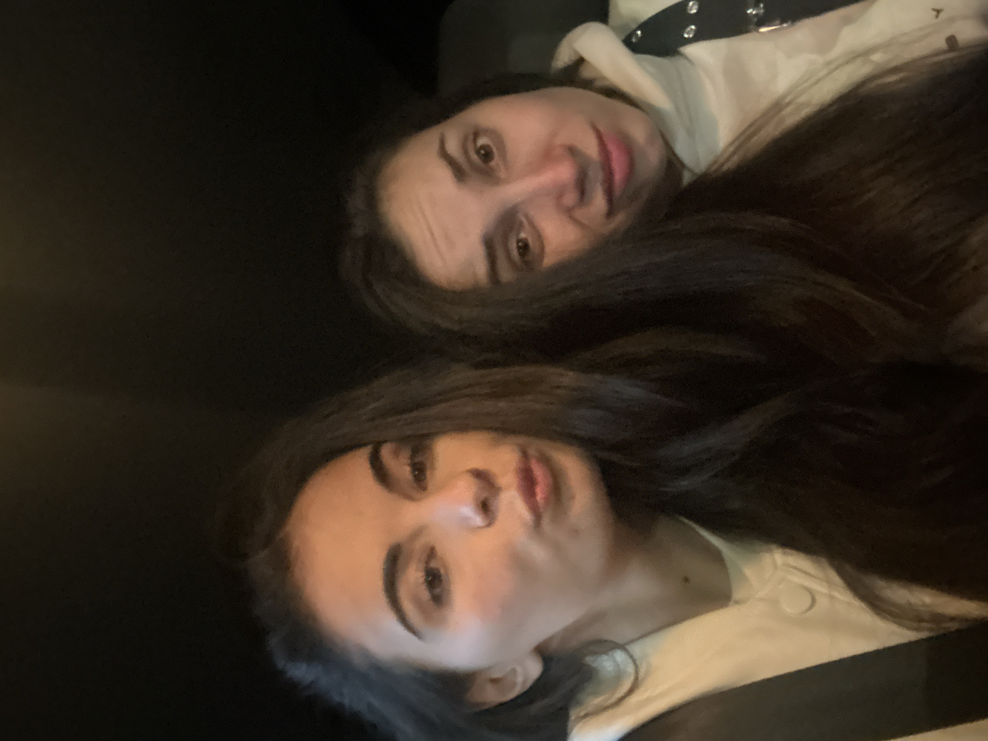 This is a close-up, vertically oriented selfie taken by two people who appear to be lying down. The person in the foreground is a woman with long, dark hair, light skin, and is making a kissing face. Her eyes are closed and her lips are puckered. She is wearing a white jacket. The second person, a woman, is in the background. She has long, dark hair and is looking at the camera with her mouth slightly open. She is wearing a white hoodie with a black strap across the chest. The lighting is dim, suggesting it could be nighttime or an indoor setting. The background is dark and out of focus. No text is visible in the image.