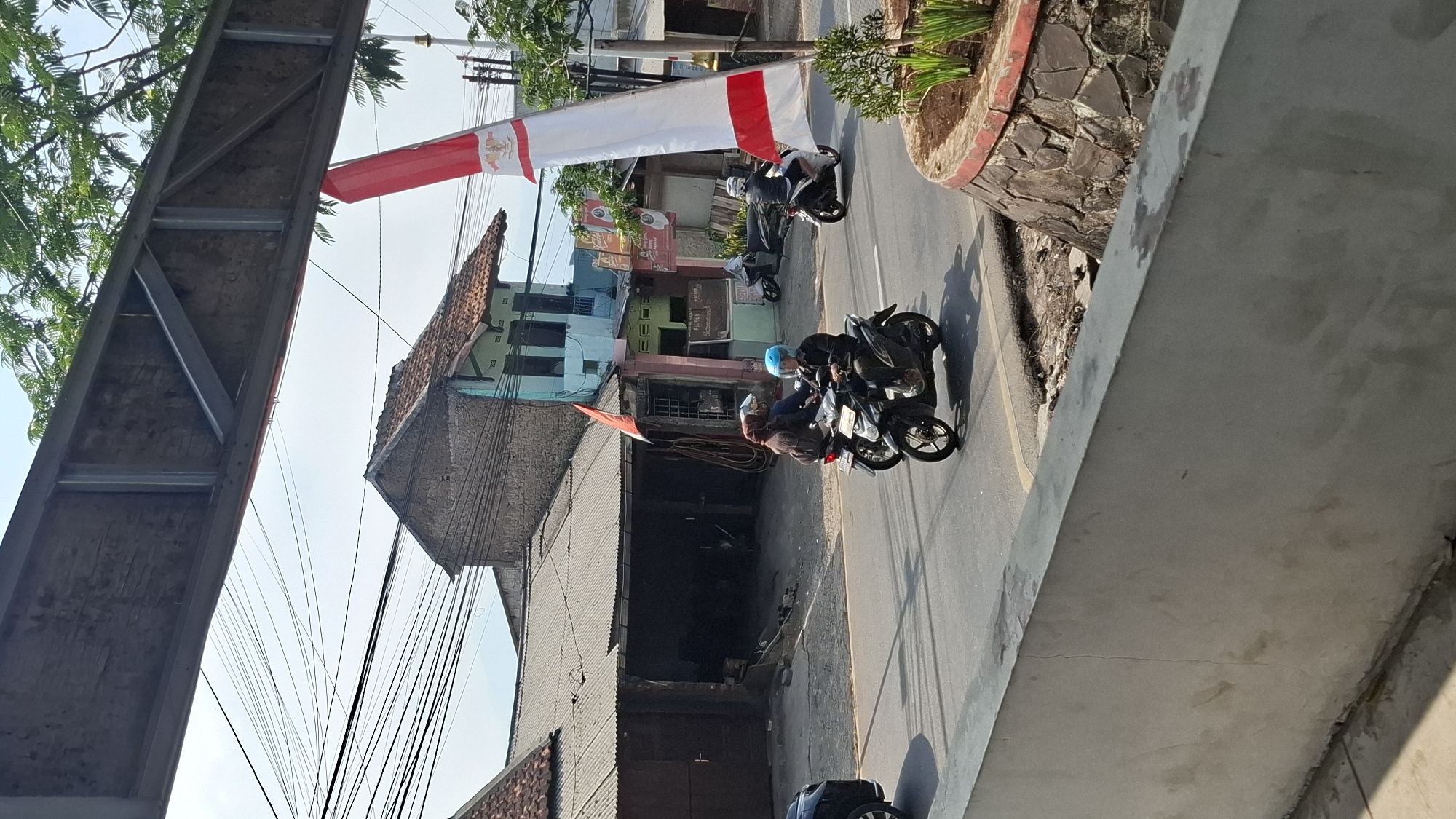 The image captures a street scene in Cilaku, Indonesia, with a focus on vehicles and buildings under a bright sky. In the foreground, a light-colored concrete wall runs horizontally across the bottom of the frame. Behind this wall, a paved road is visible, with two motorcycles in motion. The first motorcycle, black and silver, is ridden by a person wearing a blue helmet and a dark jacket, heading towards the right side of the image. Another motorcycle, also black, is positioned slightly behind and to the left, with its rider wearing a brown jacket and a light-colored helmet, also moving towards the right. Further down the road, a third motorcycle, black with white accents, is seen from the side, with its rider wearing a white helmet and a dark jacket, also traveling in the same direction.
On the left side of the road, there is a building with a dark, corrugated metal roof and a large, open doorway, suggesting it might be a workshop or garage. To the right of this building, a two-story structure with a light blue facade and a terracotta-tiled roof is visible. A red and white Indonesian flag is prominently displayed, attached to a pole and unfurled, leaning towards the right side of the frame, with a smaller red flag also visible on a pole near the blue building. Trees with green foliage are present in the background, and electrical wires crisscross the sky. The overall environment appears to be a mix of residential and commercial structures along a public road.