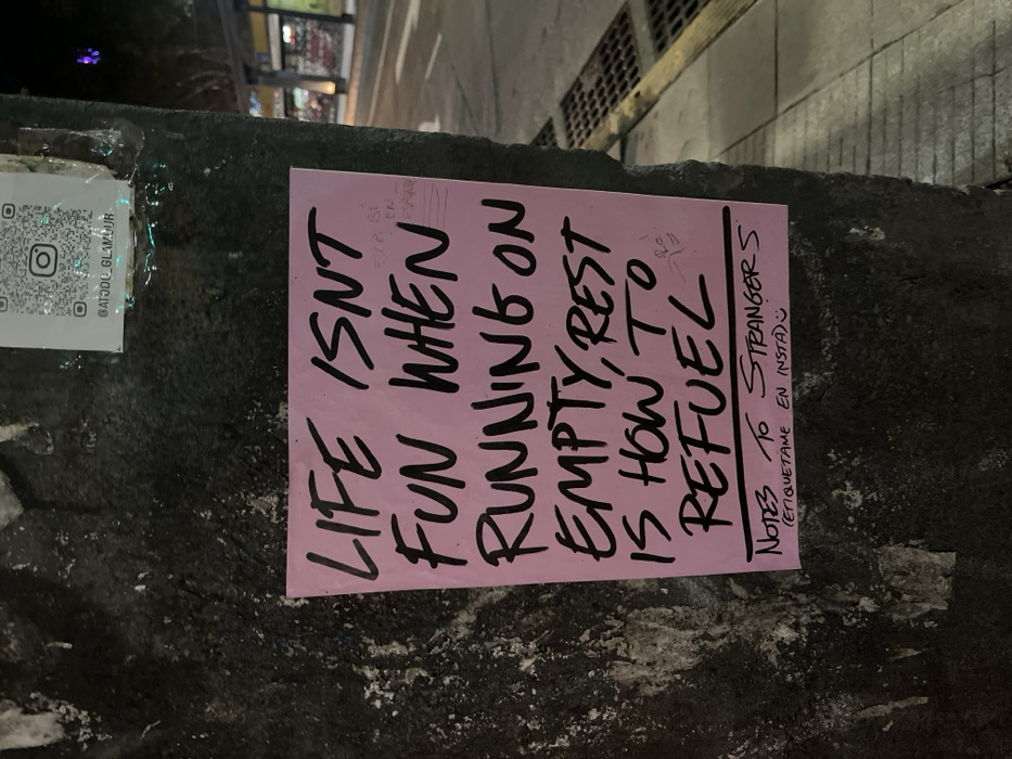 A pink flyer is plastered on a textured, dark surface alongside a QR code sticker for "GATODO GLAMOUR." The flyer's bold, handwritten text reads: "LIFE ISN'T FUN WHEN RUNNING ON EMPTY, REST IS HOW TO REFUEL." Below the message, smaller text says "NOTES TO STRANGERS (ETIQUETAME EN INSTA)." The scene appears to be an urban environment at night.