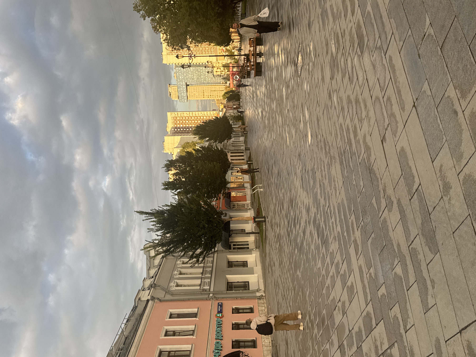 The image depicts a street scene in Moscow, Russia, likely a pedestrianized area given the paving and lack of cars. In the foreground, a person with a backpack stands facing away from the camera, on a wet, tiled pavement. To the left, a row of buildings features pastel-colored facades, with windows and architectural details characteristic of historic European city centers. Further down the street, trees are planted along the walkway, and benches are visible. In the distance, modern, tall buildings with apartment blocks are prominent, suggesting a mix of old and new architecture. The sky is cloudy, and the lighting indicates either early morning or late afternoon. Several other individuals are walking in the distance.