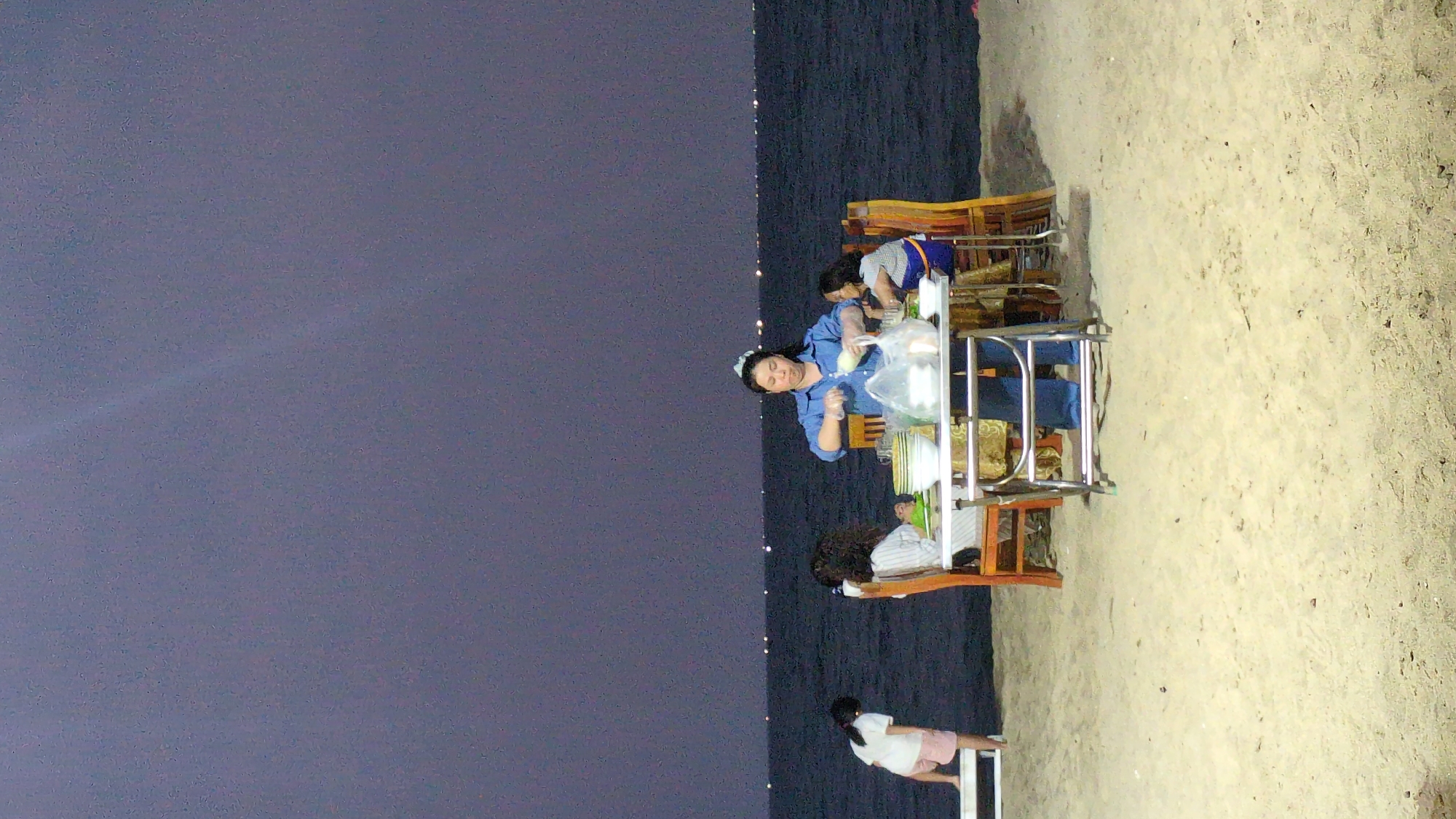 A beach scene at night in Khánh Hải, Vietnam, shows a woman in a blue shirt preparing food at a mobile food cart. Two individuals are seated nearby, presumably customers. A child stands to the side of the cart. The scene takes place on a sandy beach with the ocean visible in the background. The lighting indicates an evening or nighttime setting.