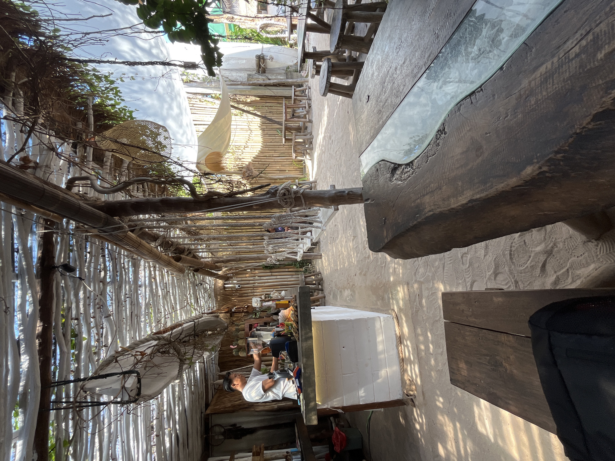 The image depicts an open-air cafe with a thatched roof and sand flooring, likely in Karimunjawa, Indonesia, given the tropical ambiance and common cafe style in the region. A man in a white t-shirt is preparing drinks behind a white brick counter, suggesting he is a barista. The cafe features rustic wooden tables and stools, with swings hanging from the ceiling, creating a relaxed and bohemian atmosphere. The background is adorned with bamboo walls and lush greenery, indicating an outdoor setting. The lighting suggests it is daytime, with sunlight filtering through the roof, casting shadows on the sand.