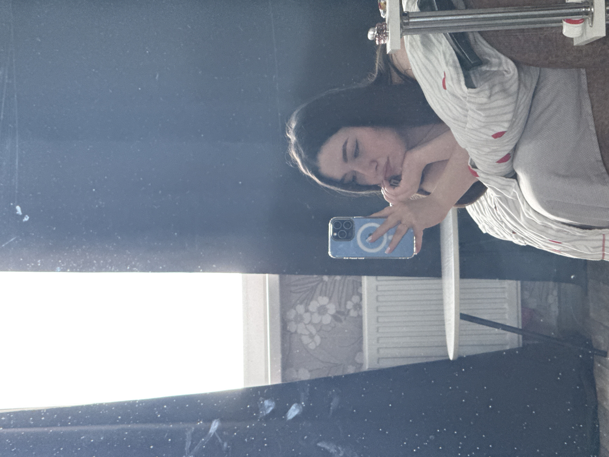 A mirror selfie shows a young woman in a light-colored striped shirt. She's holding a blue smartphone with a prominent camera array. The background reflected in the mirror includes a window providing significant light, a wall with floral wallpaper, and a white radiator. A portion of what seems like photo equipment can be seen above the woman's head. There is no data on the location in Perm, Russia, nor additional information.