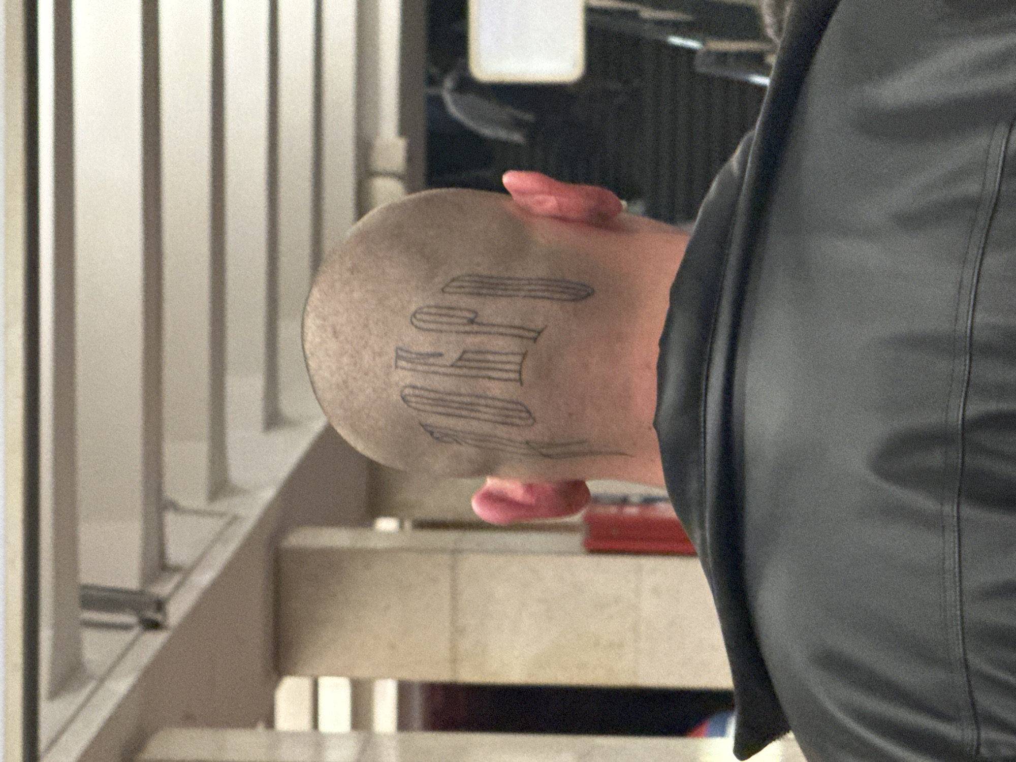 A close-up shot focuses on the back of a person's head, which is shaved and features a tattoo in black ink of what appears to be Gothic-style lettering. The tattoo appears to read "EL". The subject is wearing a black leather jacket or vest. The background includes a staircase with white railings and beige tiles. The setting suggests an interior location, potentially a public space like a metro station. There is not enough information to identify the location in Moscow, Russia.