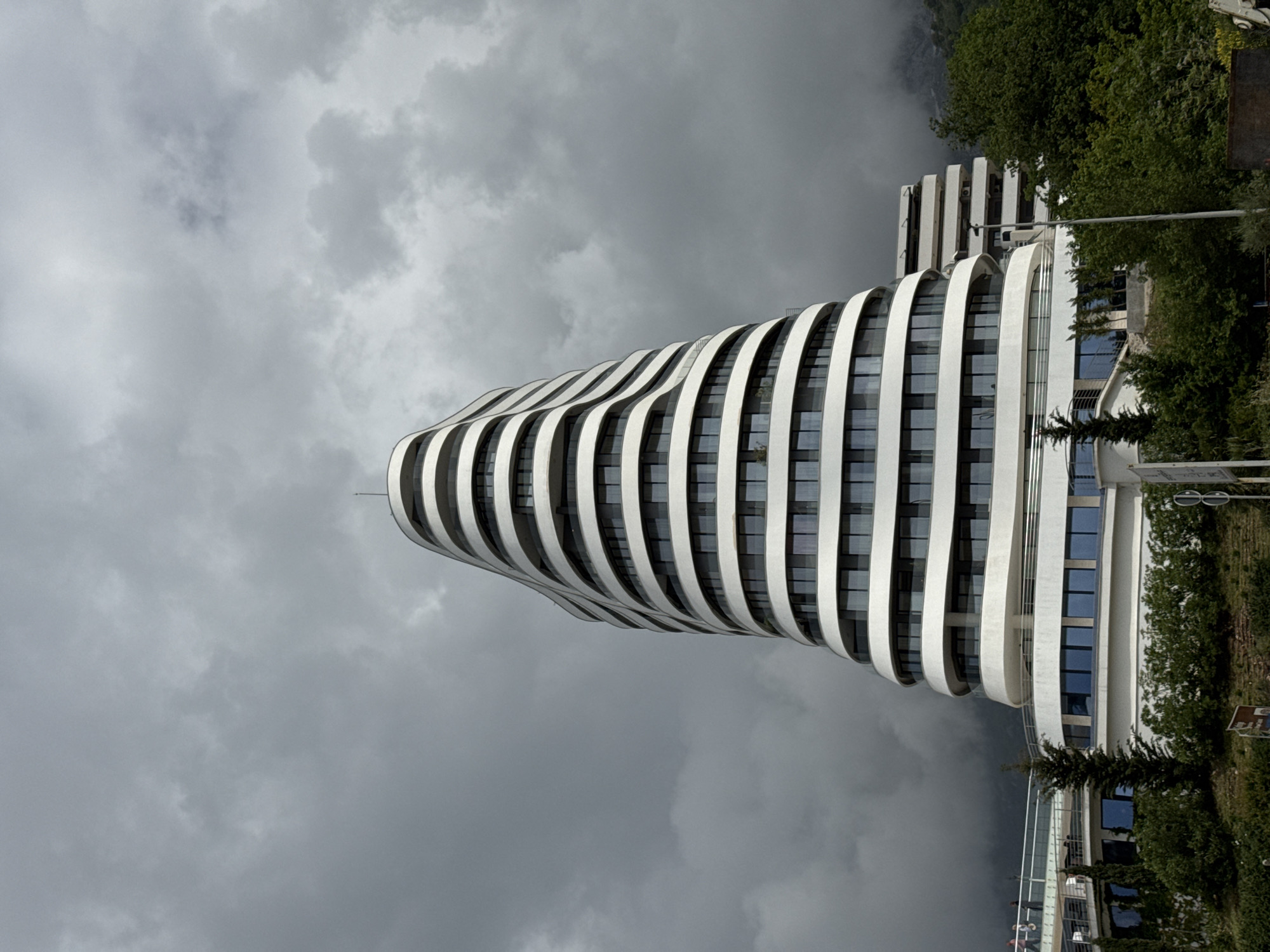 A ground-level, full outdoor shot depicts a uniquely designed, modern building situated in Boreti, Montenegro, under a heavily clouded sky. The structure features a curvilinear design with alternating white architectural bands and windows. The building appears to rise out of a landscaped area with trees and some street furniture present. Other sections of the building are visible beyond the main curved section, including what seems to be a traditional tower shape. The weather looks overcast.