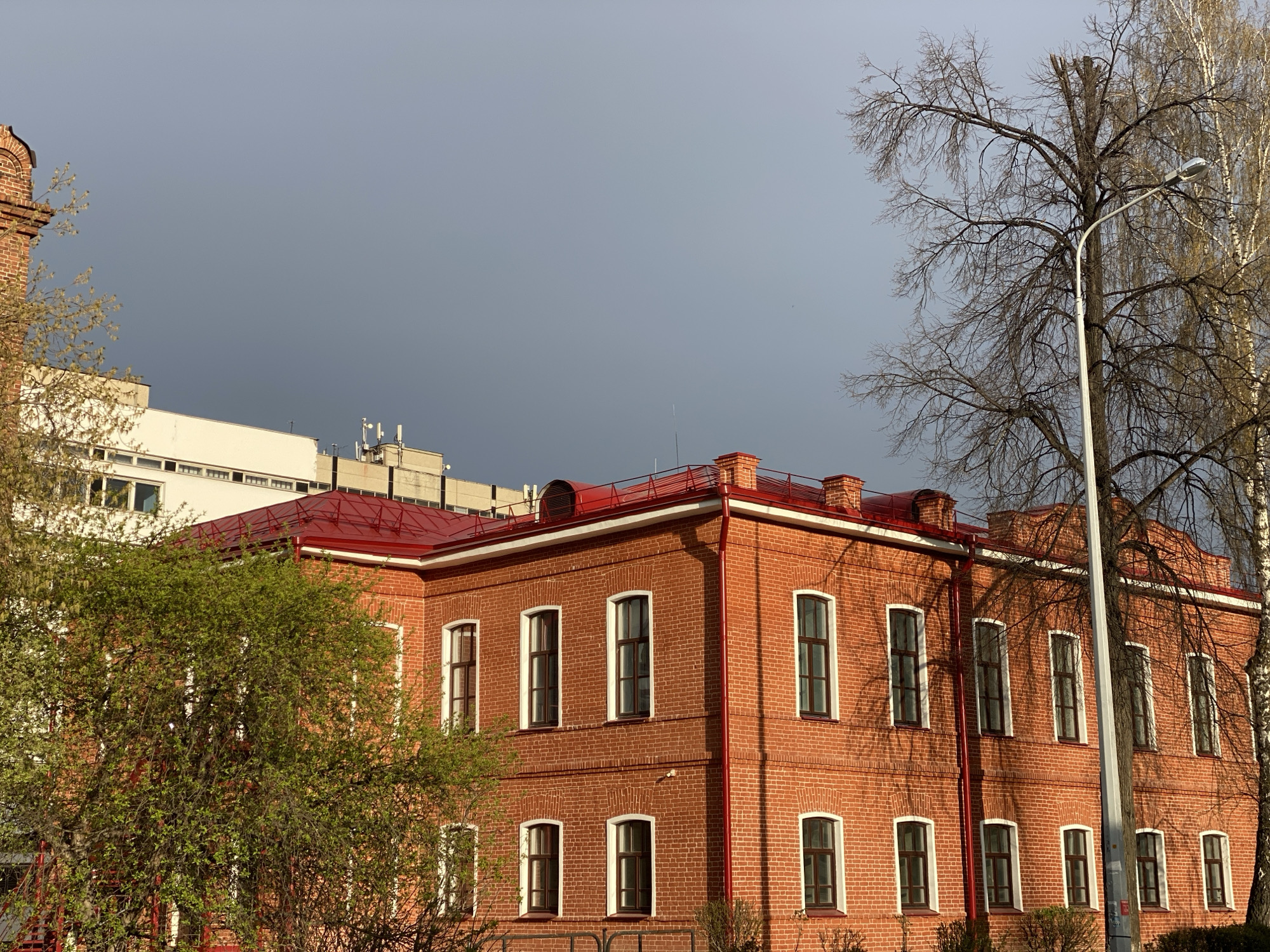 The image presents an urban landscape in Yekaterinburg, Russia, dominated by a two-story red brick building with a vivid red roof. The building exhibits classic architectural elements, including arched tops on its numerous rectangular windows, which feature white frames and dark sashes. Direct sunlight illuminates the brickwork and the bright red roof, casting defined shadows on the building's facade, particularly from the branches of a large, bare tree to the right.
To the left of the brick building, a lush green tree with fresh spring foliage partially obscures the view. Behind the main building, parts of modern, lighter-colored buildings with antenna installations are visible. On the right, a tall, bare deciduous tree with a dense network of branches, possibly with a broken top, stands prominently, accompanied by a slender, lighter-barked tree further back, likely a birch. A modern silver street light pole rises vertically in front of the bare tree.
The sky is a notable element, displaying a dramatic contrast. While a small section in the upper left shows a clearer, lighter sky, the majority of the overhead expanse is a dark, overcast grey, suggesting either an approaching storm or recent heavy clouds. The strong, directional sunlight on the building and the green tree, coupled with the bare trees and budding foliage, indicates early spring. There are no people visible, and no specific activities or visible text can be discerned. The scene captures a moment of striking architectural presence against a moody, transitional sky.