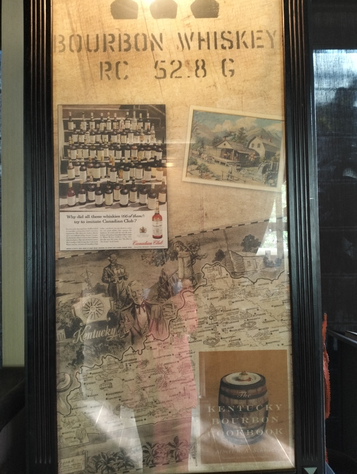 A framed display features a collection of images and text related to Kentucky bourbon whiskey.
The top section displays the stenciled words "BOURBON WHISKEY" with "RC 52.8 G" underneath. Below this, a vintage Canadian Club advertisement shows a display of whisky bottles with the headline "Why did all these whiskies (66 of them!) try to imitate Canadian Club?" A print of a rural landscape scene is adjacent to the ad.
A map of Kentucky occupies a significant portion of the display, showing city names like Lexington and Louisville. Above the map, sketched figures include a man in a suit and a man holding a tray. At the bottom, an image of "The Kentucky Bourbon Cookbook" with a barrel on the cover completes the collage.
The frame is dark brown and consists of multiple vertical grooves. The background is a textured tan color, which is reminiscent of a wooden plank.
The location is not specified or identified in the image.