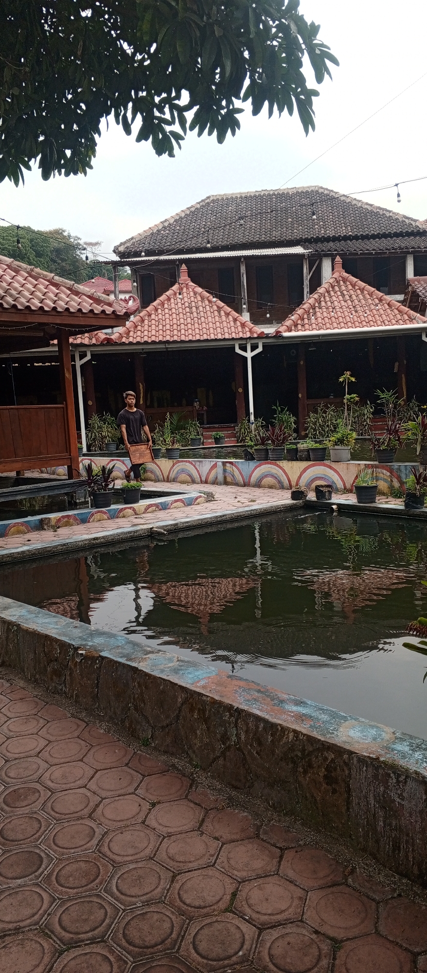 In Plaosan, Indonesia, a young man walks along a tiled path next to a pond. He carries a wooden tray. Behind him, a building with a tiled roof features open-air seating areas with potted plants. The sky is overcast.