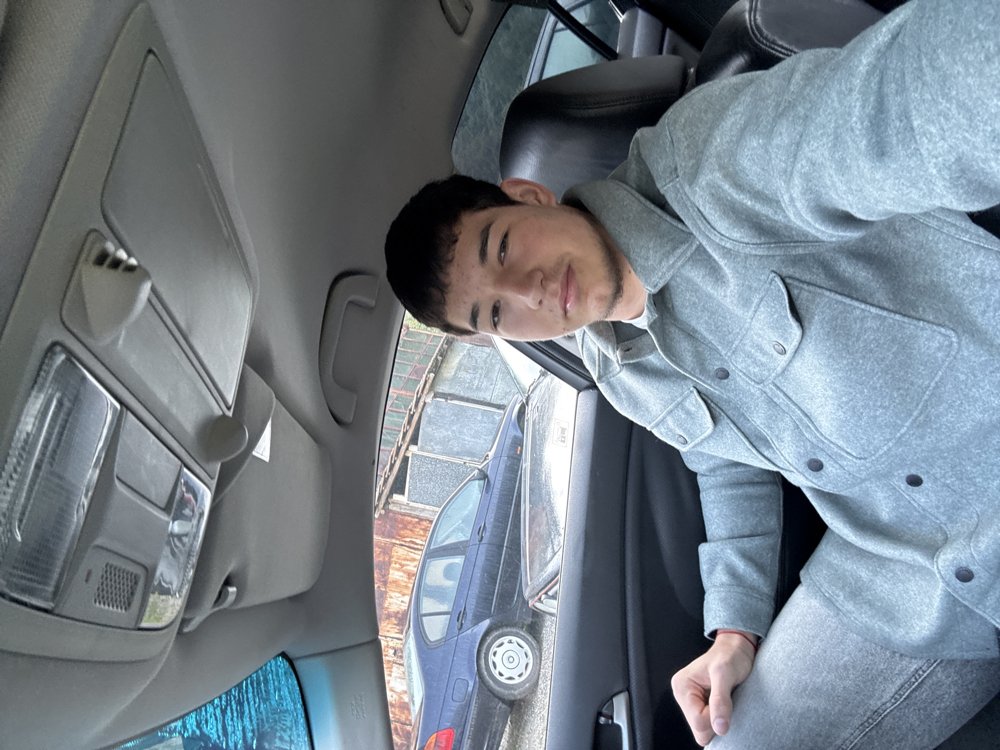 A young man sits in the driver's seat of a car, possibly parked. He is wearing a grey button-down shirt or jacket and matching grey pants. He faces the camera. The car is parked next to another car and near a rusted, metal structure, possibly part of a building. The interior of the car includes overhead lights and a sun visor. The location is potentially Zlatograd, Bulgaria, based on the prompt's requirement.