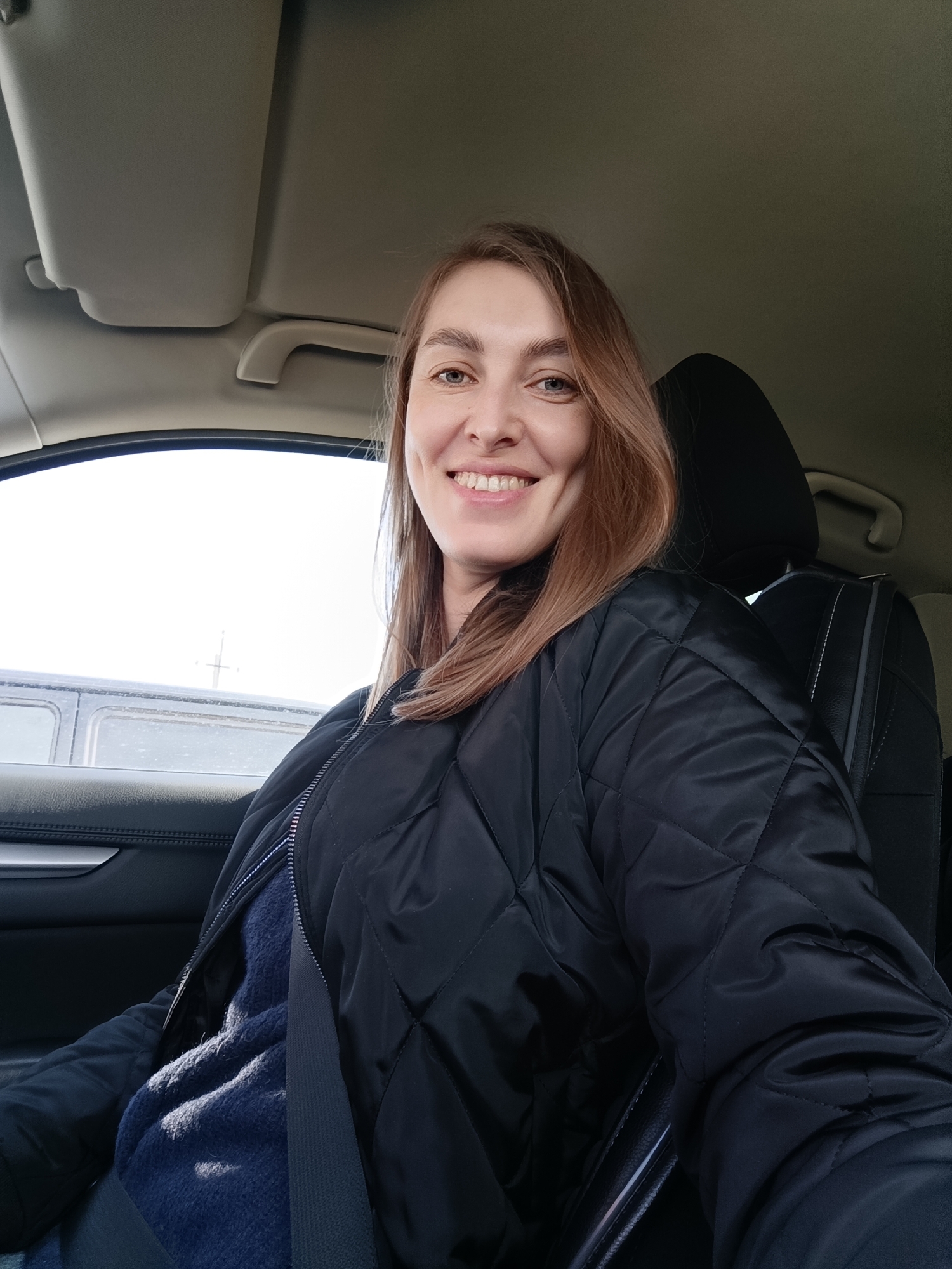 A woman is seated in the driver's seat of a car. She is wearing a black quilted jacket over a blue sweater and has her seatbelt fastened. The interior of the car is visible, including the dashboard and headrest. Outside the car window, a grey barrier or structure can be seen, likely a bridge. There is no information available to pinpoint the precise location as Chaikovsky, Russia from this image alone.