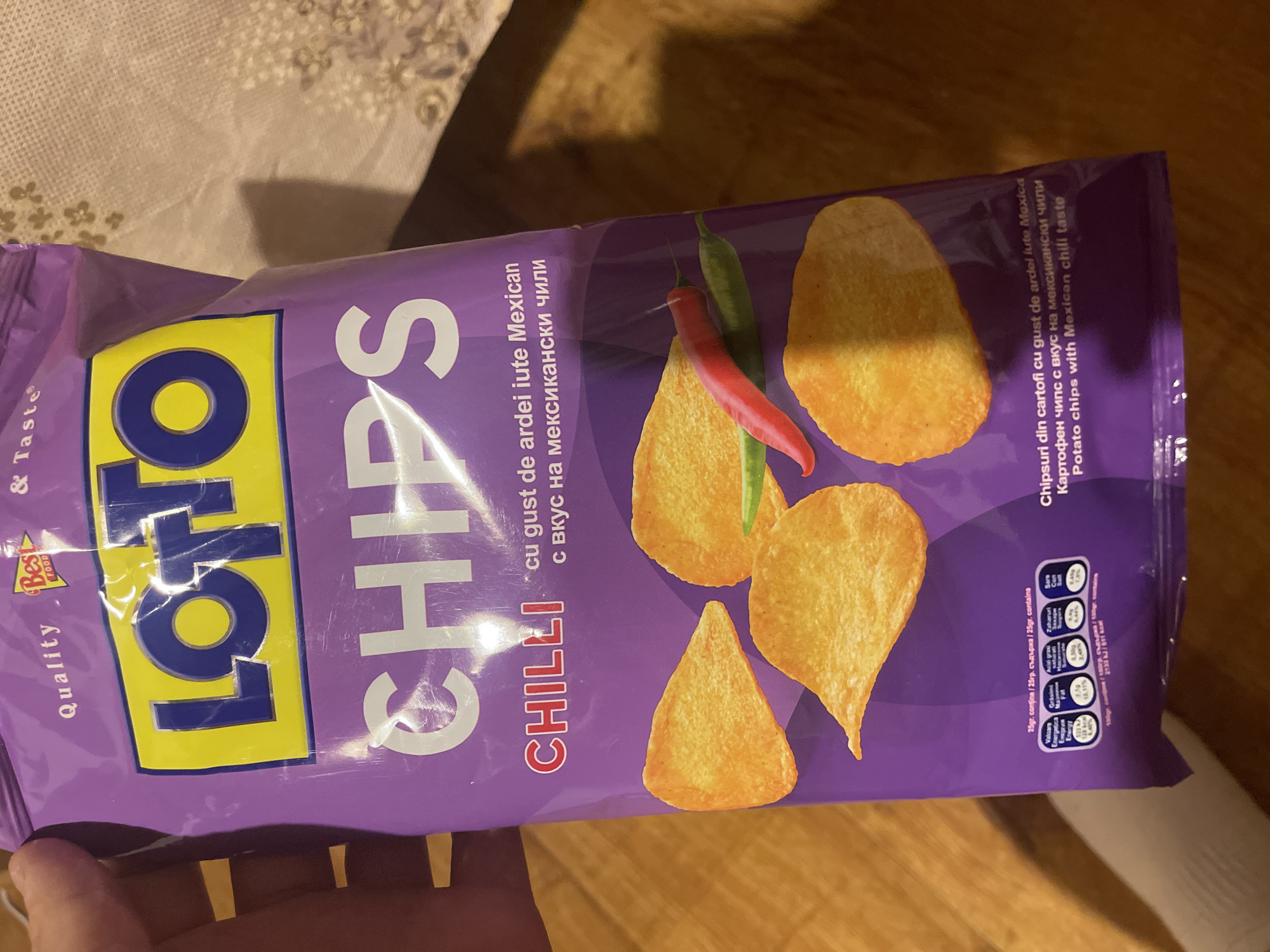The image captures a purple bag of "LOTTO CHIPS CHILLI" potato chips, held by a hand, against a light-colored patterned surface, possibly a tablecloth. The bag is positioned diagonally, showcasing the brand name "LOTTO" in yellow and blue, and "CHIPS" in white lettering. The word "CHILLI" is written vertically in red.
The flavor is described as "cu gust de ardei iute Mexican" (with Mexican chili taste) in Romanian, "с вкус на мексикански чили" in Bulgarian, and "Potato chips with Mexican chili taste" in English. The packaging design features several potato chips along with a red and a green chili pepper, illustrating the flavor profile. A "Quality & Taste" label with a "Best FOOD" logo is visible on the upper left.
Nutritional information is displayed on the right side of the bag. For a 25g serving, it states: Energy 533 kJ / 128 kcal (6.4% of Reference Intake), Fats 8.35g (11.9% RI), Saturated fatty acids 4.50g (22.5% RI), Sugars 0.44g (0.5% RI), and Salt 0.39g (6.5% RI). The energy for 100g is listed as 2133 kJ / 511 kcal. Additional text includes "Chipsuri din cartofi cu gust de ardei iute Mexica" and "Картофен чипс с вкус на мексикански чили." The scene appears to be an indoor setting with ambient lighting.
