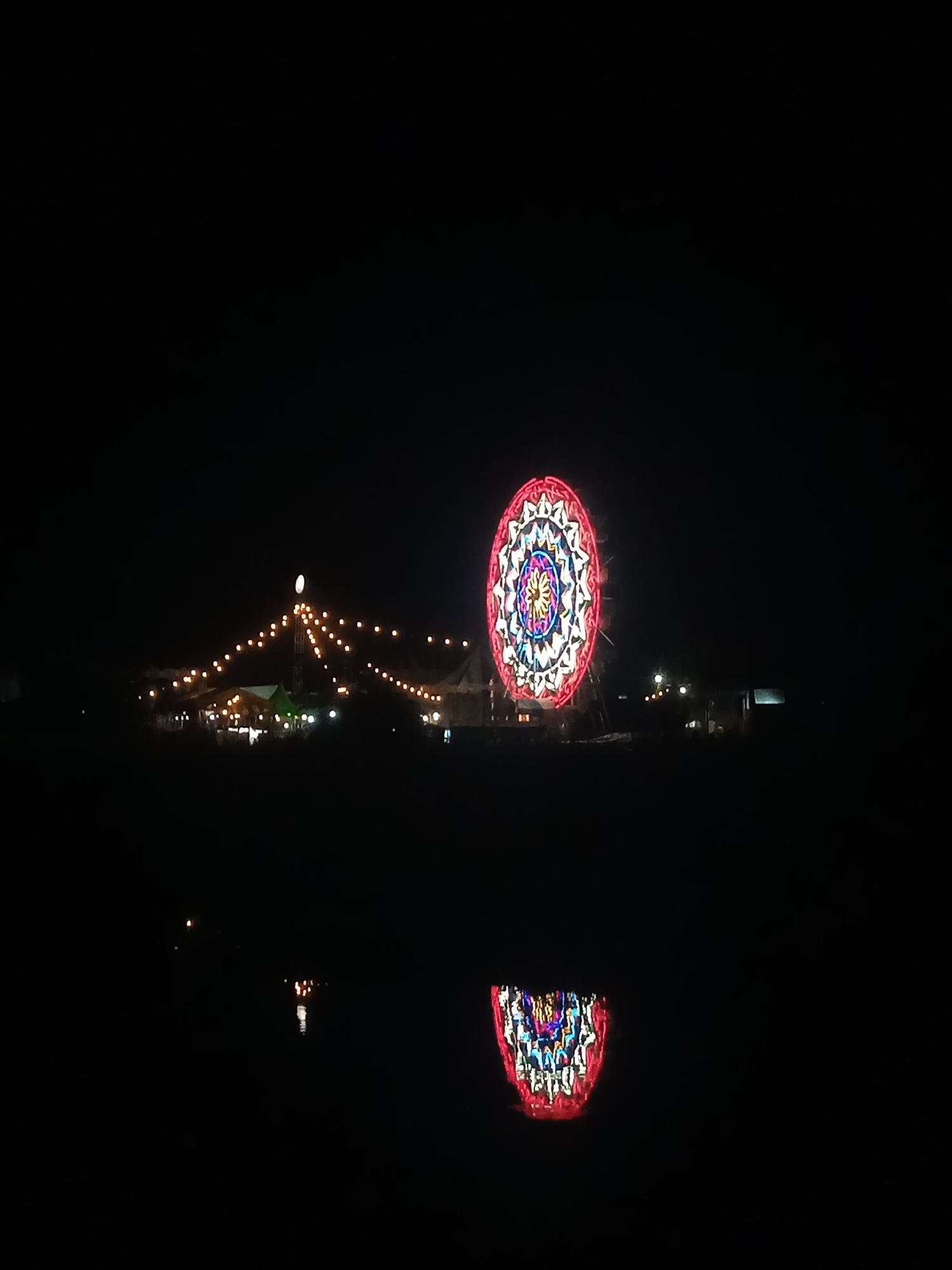A nighttime scene depicts a brightly illuminated Ferris wheel with intricate colorful patterns, dominating the right half of the frame. To the left of the Ferris wheel, a cluster of tents and structures are visible, adorned with strings of warm-toned lights. These lights stretch upwards towards a central tower, creating a festive atmosphere. The foreground consists of dark, still water, reflecting the lights of the Ferris wheel and the tents, creating a symmetrical, inverted image of the scene above. The overall impression is that of a lively night market or festival, possibly during a celebration. The image is dark with high contrast, emphasizing the illuminated elements. The location is identified as Sigli, Indonesia.